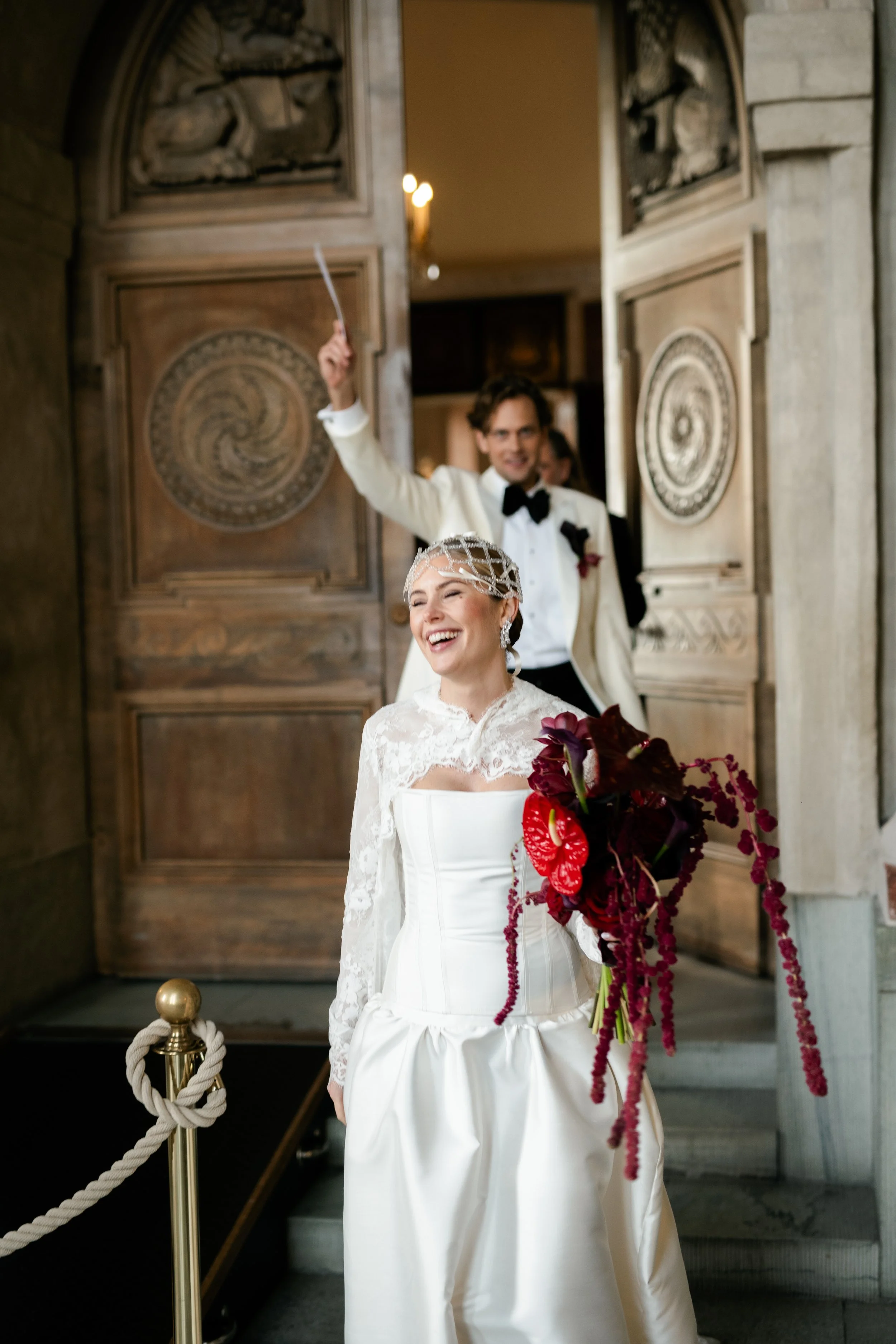 A bride and groom during a wedding ceremony, with the groom behind the bride holding a wand or stick, both smiling joyfully at the entrance of a building.
