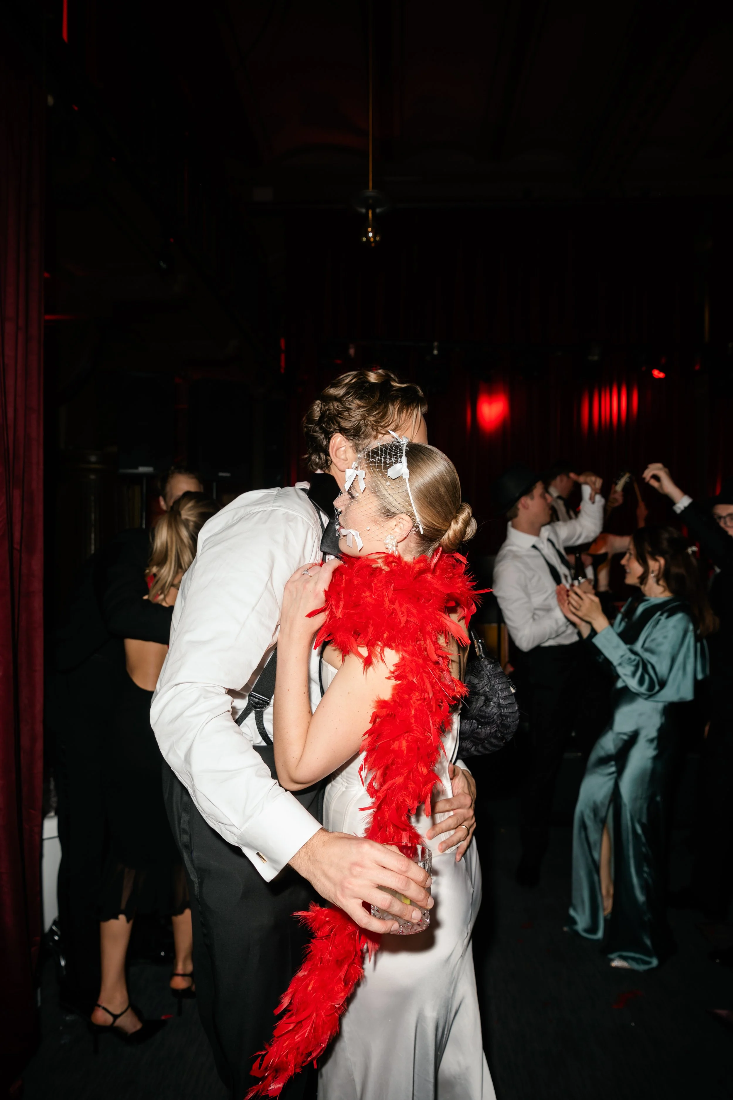 Couple dancing closely at a party. The woman wears a white satin dress and a red feather boa with a mesh face mask. The man wears a white shirt and black trousers.