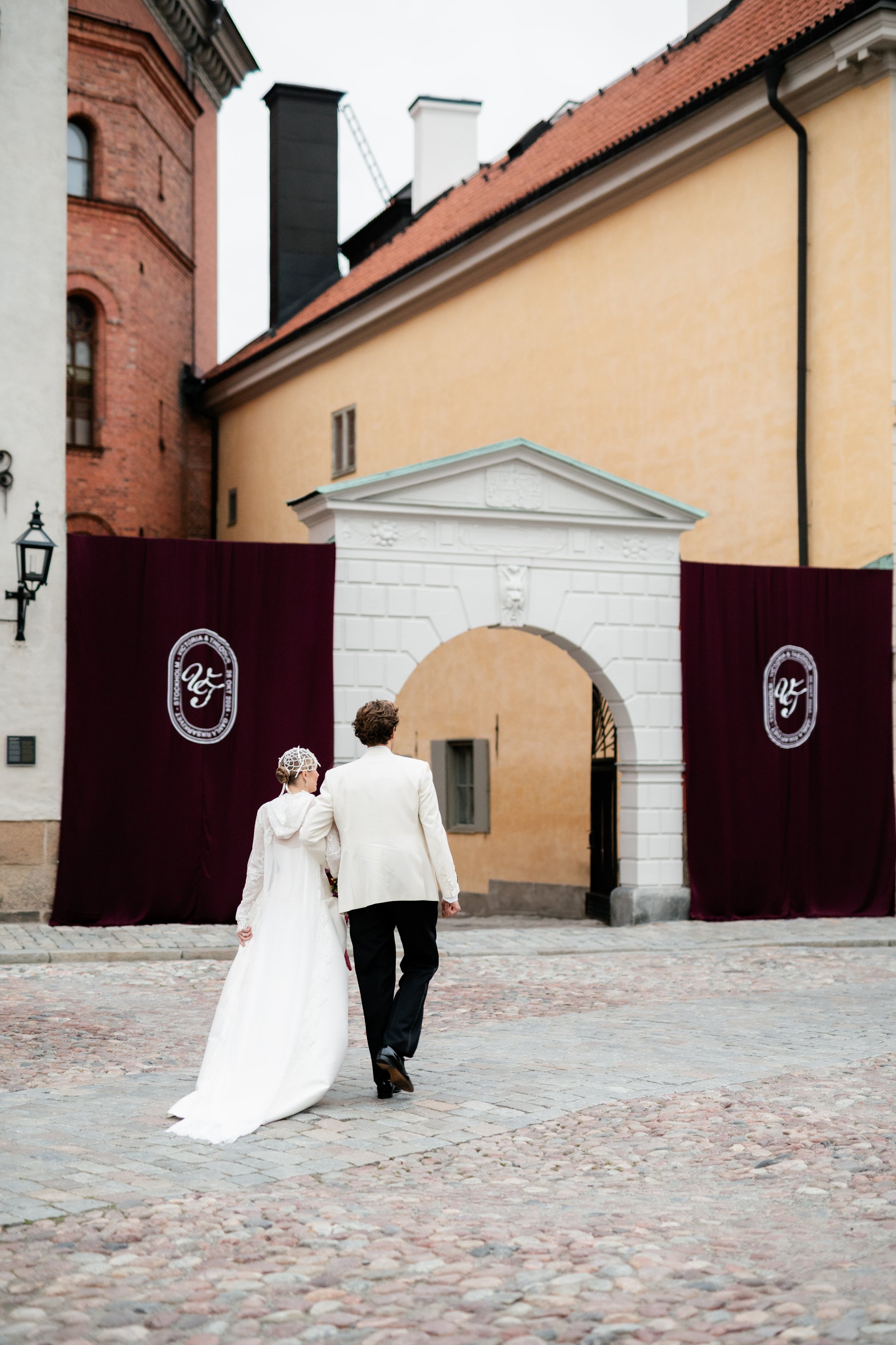 A newlywed couple, dressed in white and black, walking away from camera towards an archway with decorative banners on either side. The scene is set on a cobblestone street with historic buildings.