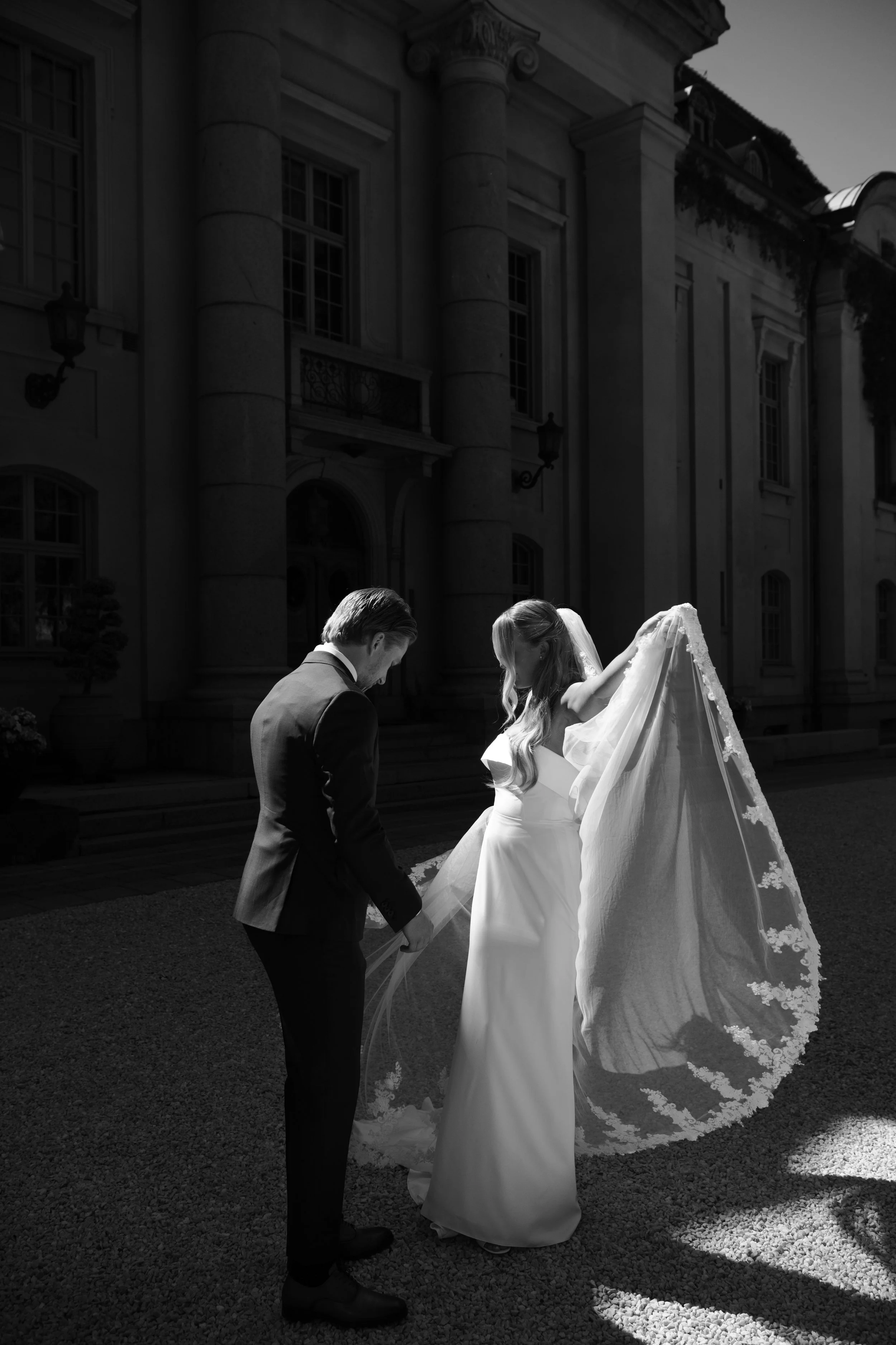 A bride and groom standing outside a large building, holding hands, with the bride lifting her veil. The scene is lit to highlight their figures, with the building in the background.