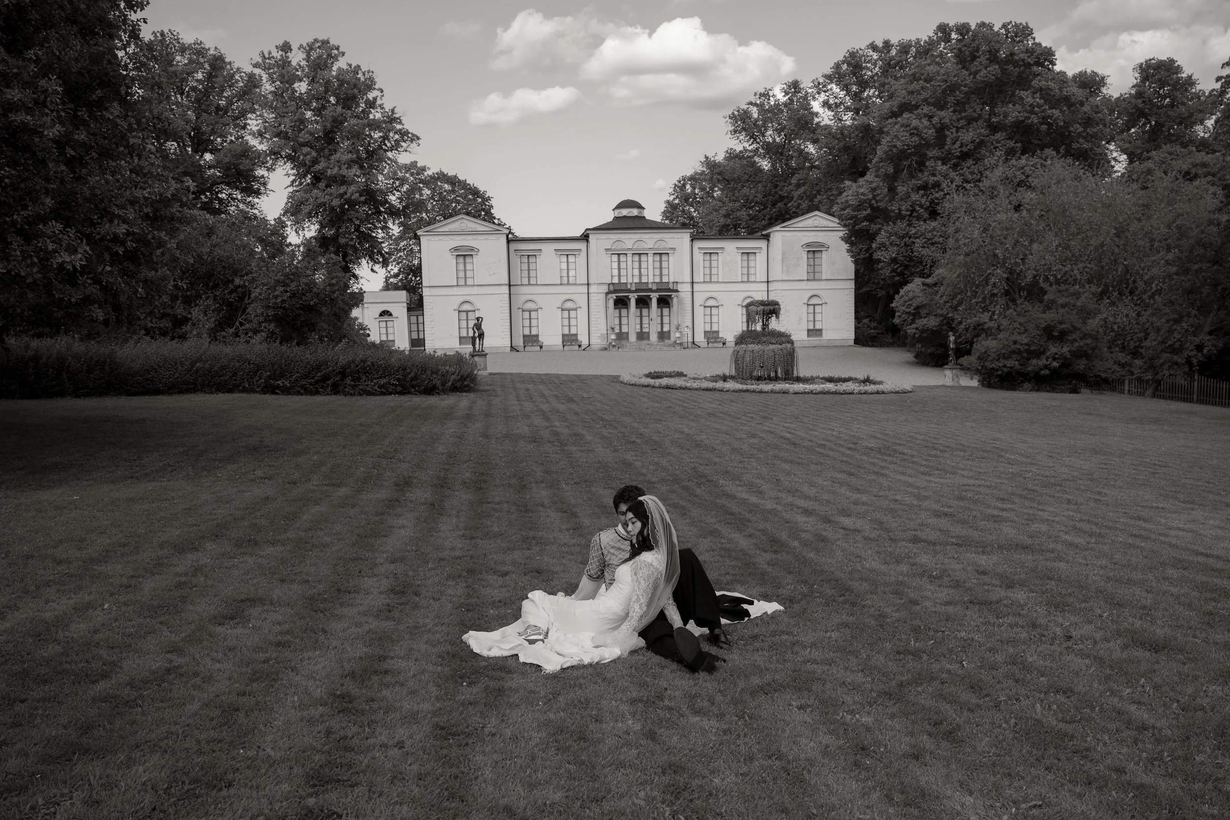 Black and white photo of a bride and groom sitting on grass in front of a large historic estate with trees and sculptures.