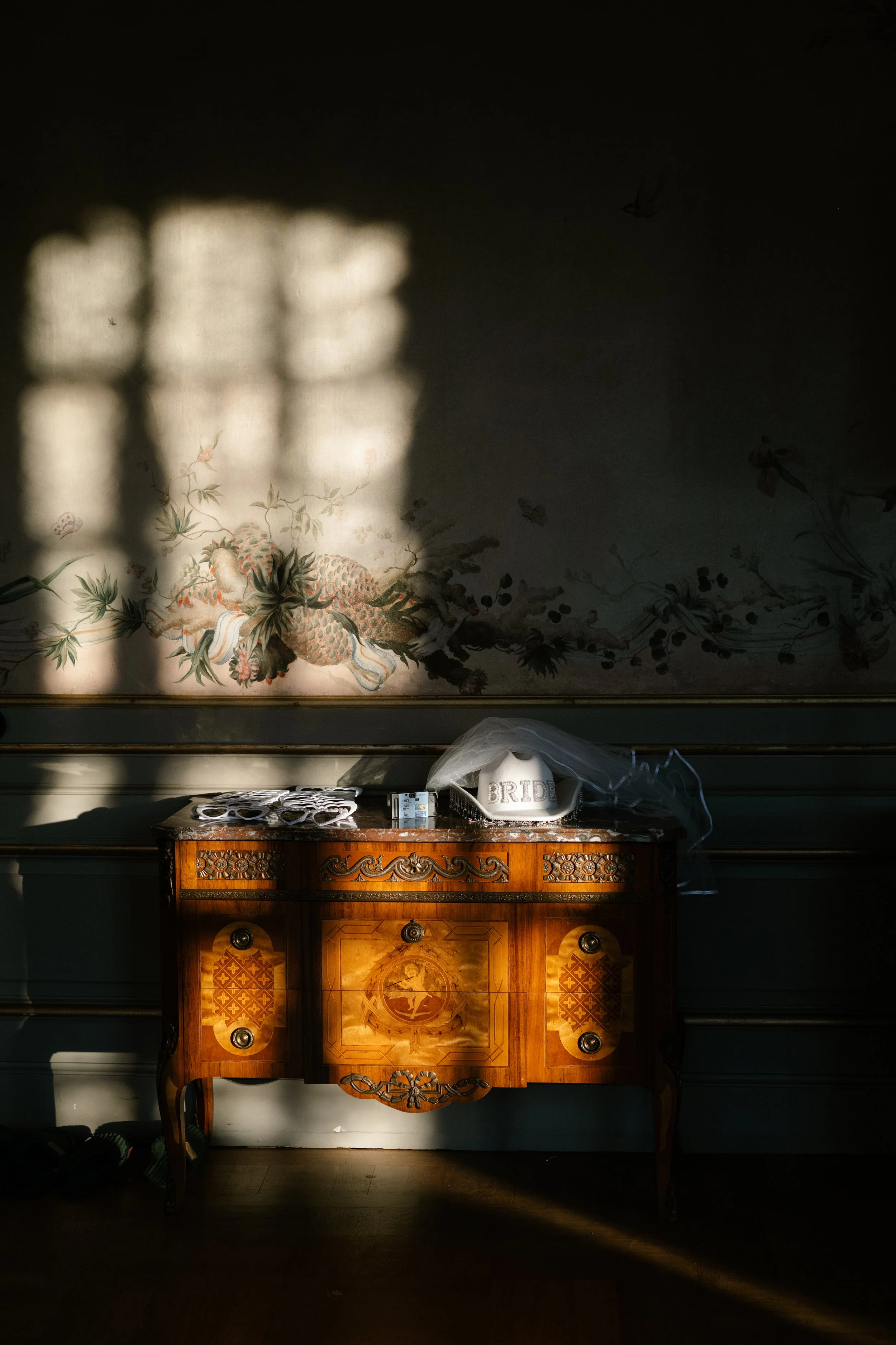Sunlight creates a window shadow on a wall above a vintage wooden dresser decorated with floral patterns. On the dresser, there are a white ceramic item with the word 'BRIDE' on it, a veil, and some small objects. The wall behind features a mural wit