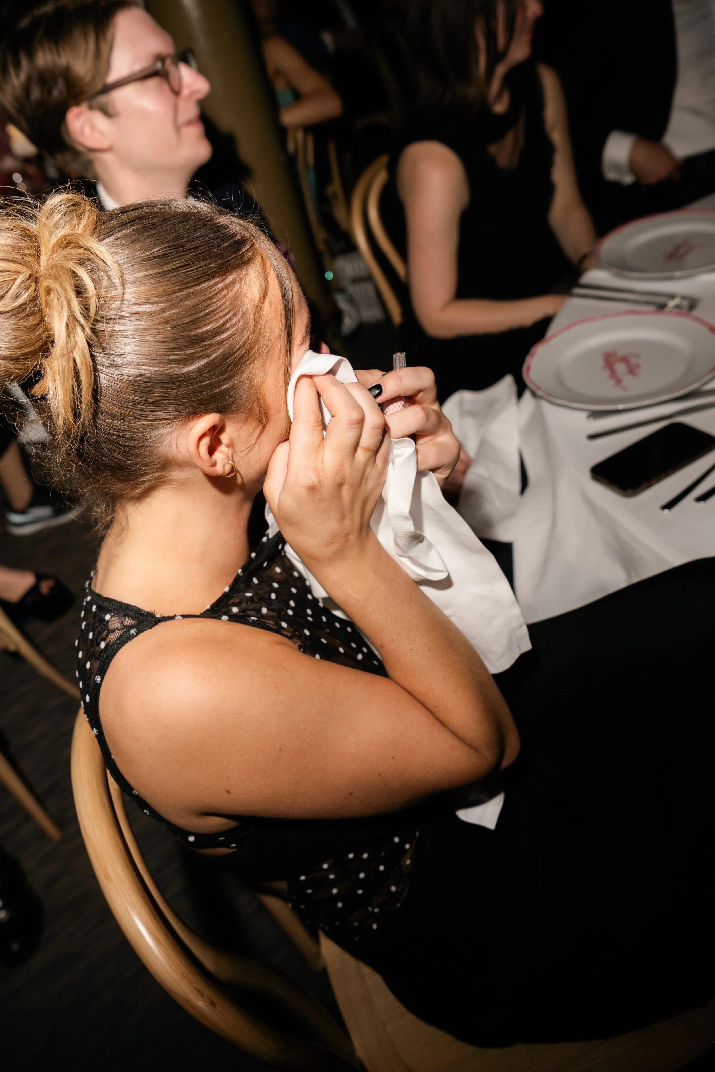 A young woman with a ponytail and black dress with white dots is crying and wiping tears with a tissue at a formal dinner event.