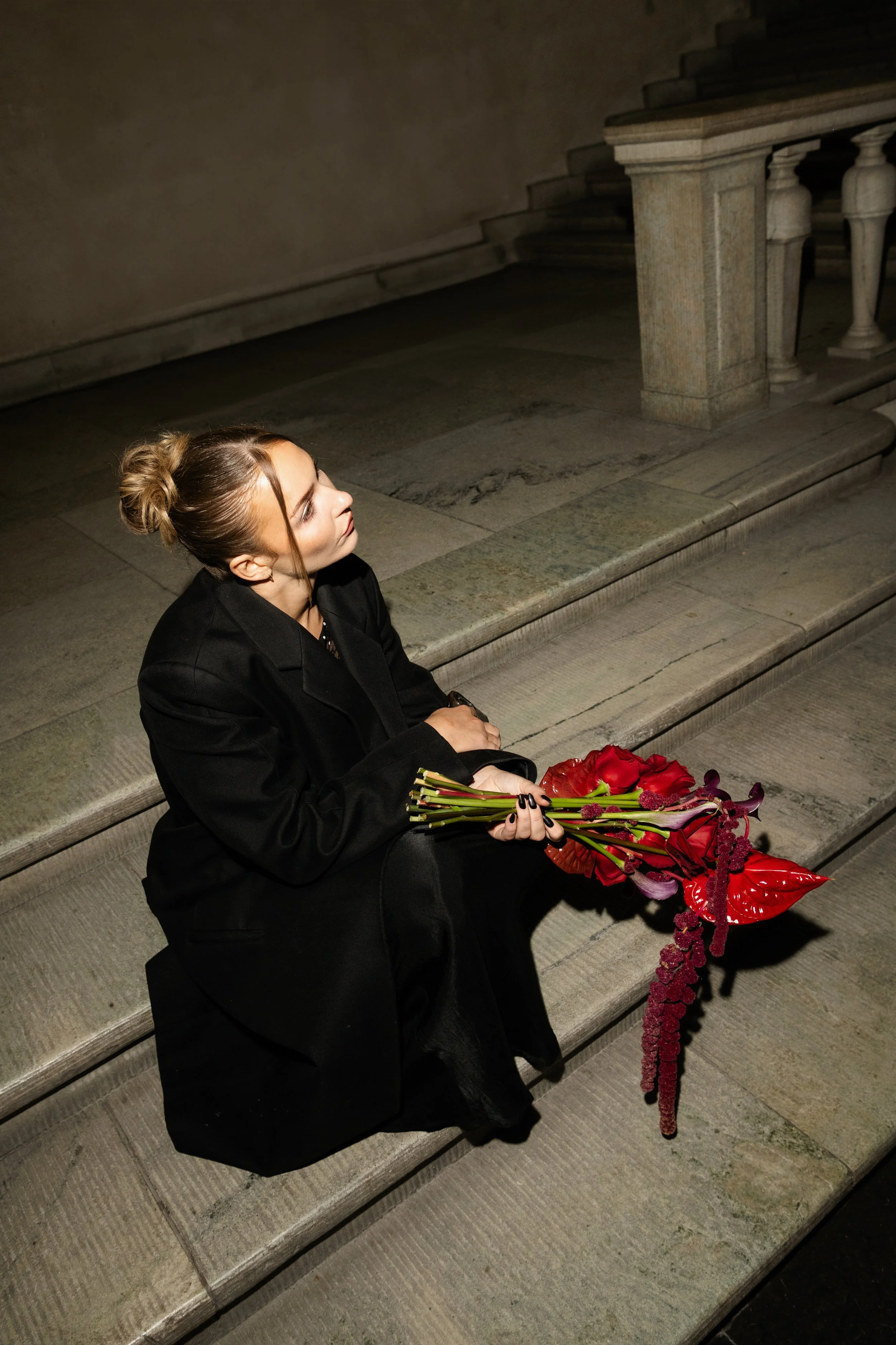 A woman with light skin and blonde hair tied in a bun, dressed in black, sitting on stone steps at night, holding a bouquet of red and purple flowers.