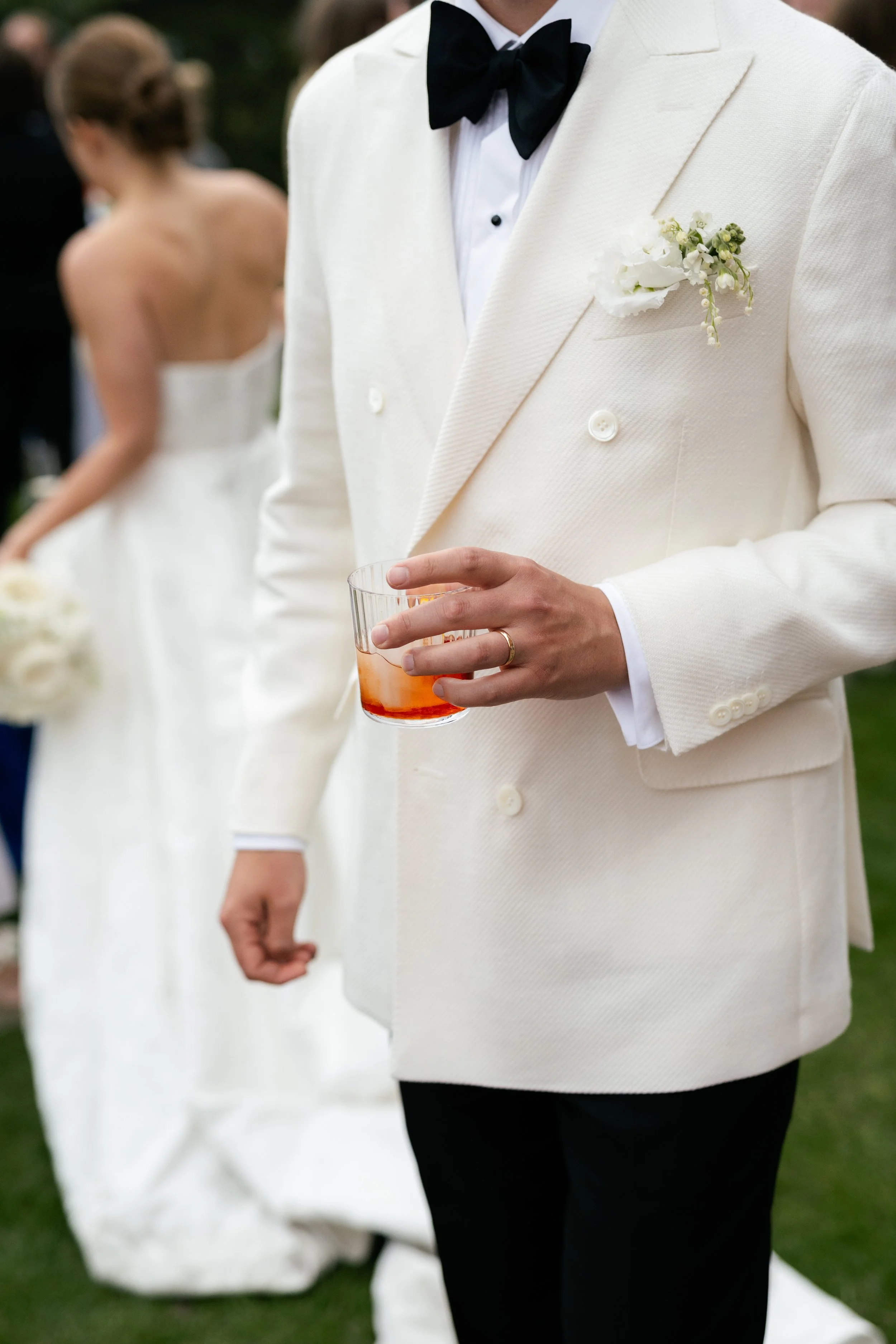A man wearing a white tuxedo with a black bow tie holding a glass of amber-colored drink at an outdoor wedding reception. A woman in a white strapless wedding gown holding a bouquet is blurred in the background.