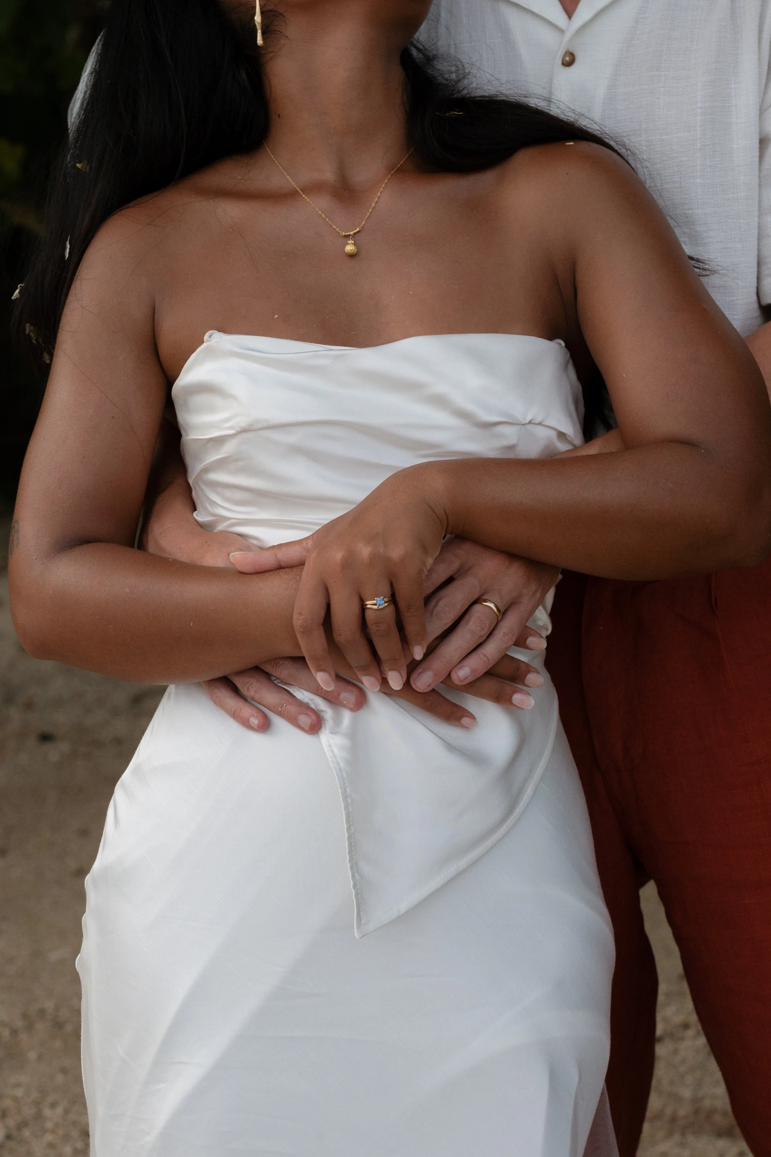 Close-up of a woman in a white satin strapless dress with jewelry, embracing a person in a light-colored shirt, against a natural outdoor background.