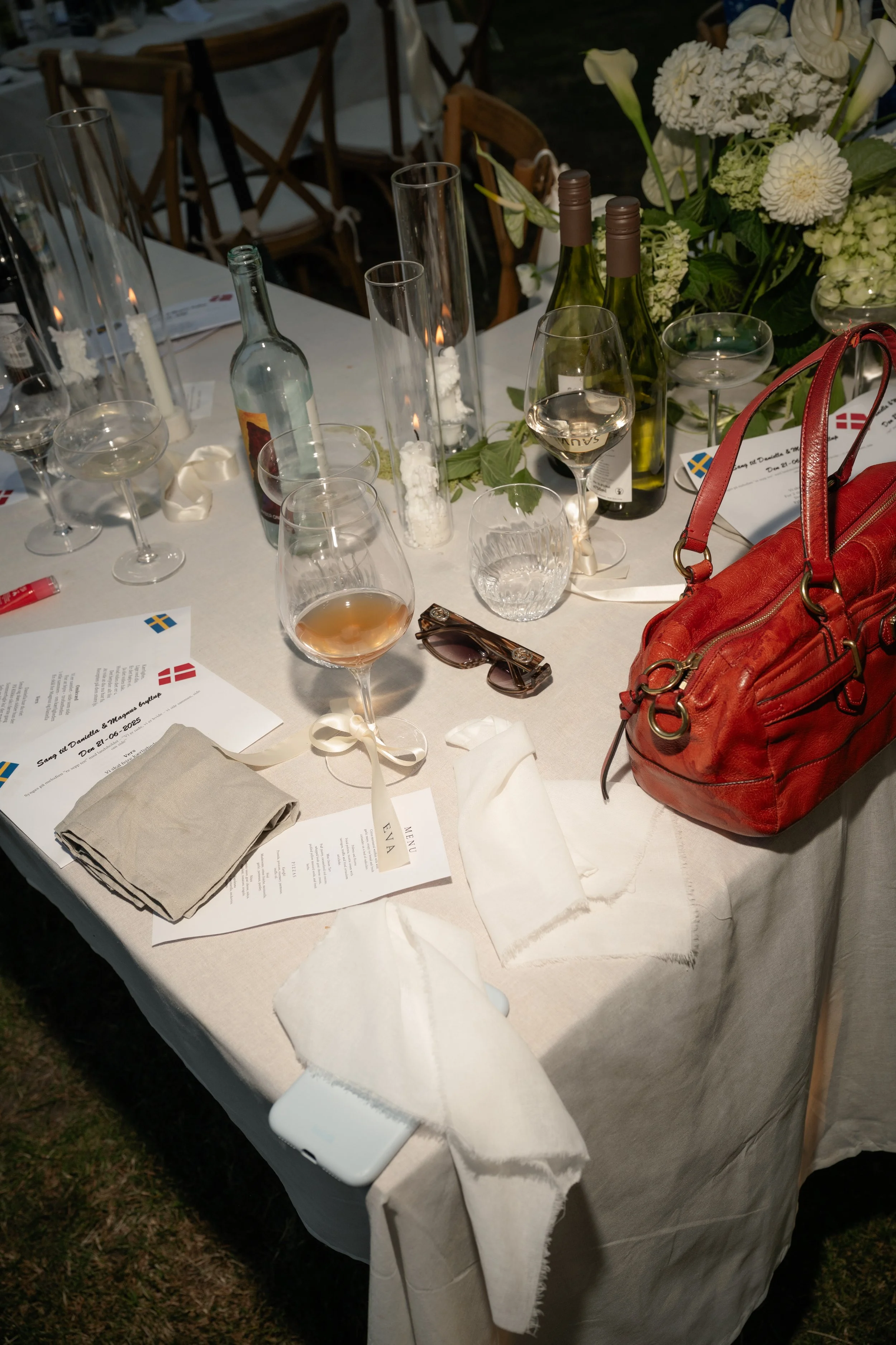 A table set for a celebration with various glasses, bottles of wine, a red purse, sunglasses, and floral decorations.
