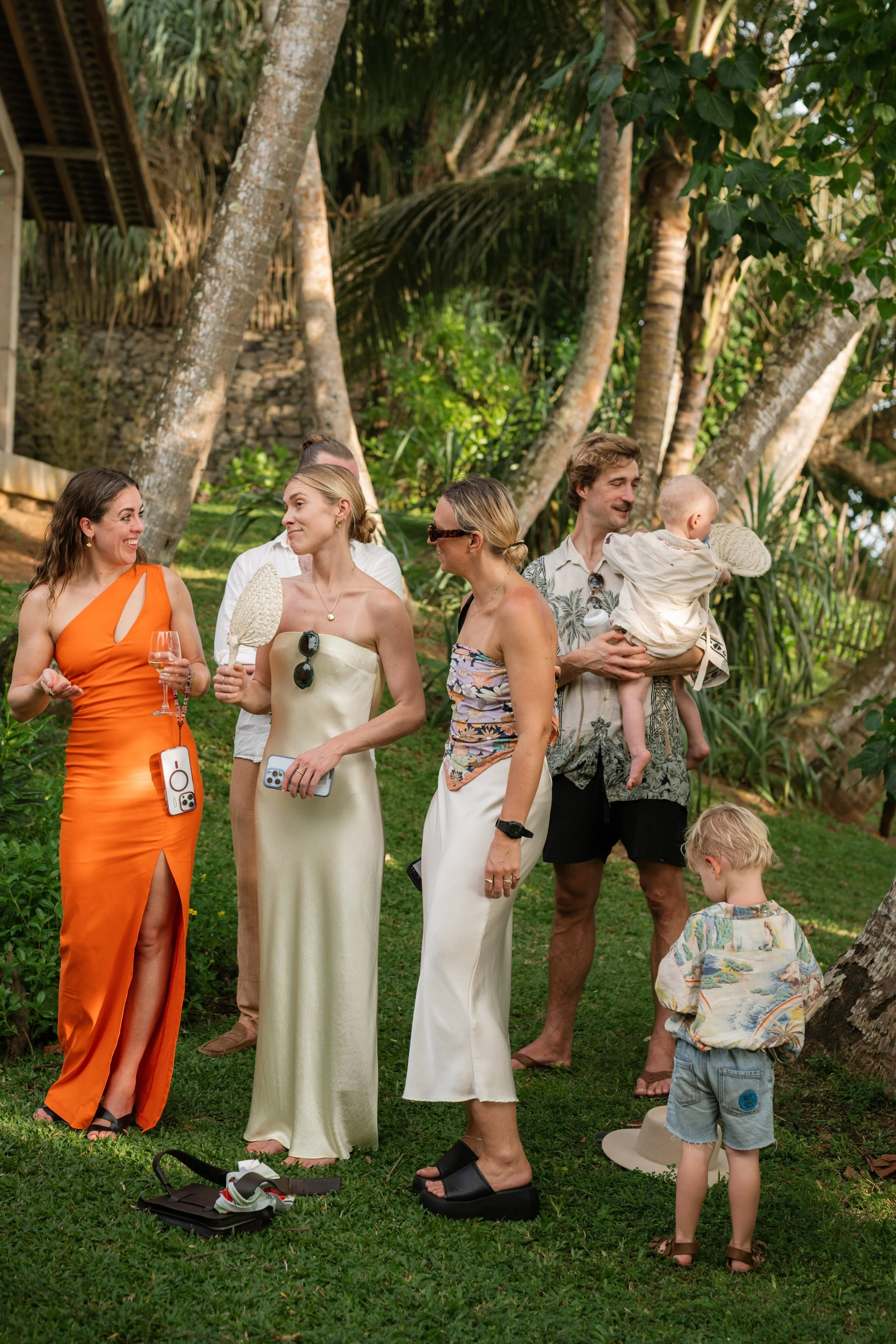 People gathered outdoors in a tropical setting, dressed in summer attire, engaging in conversation and holding drinks during a social event.
