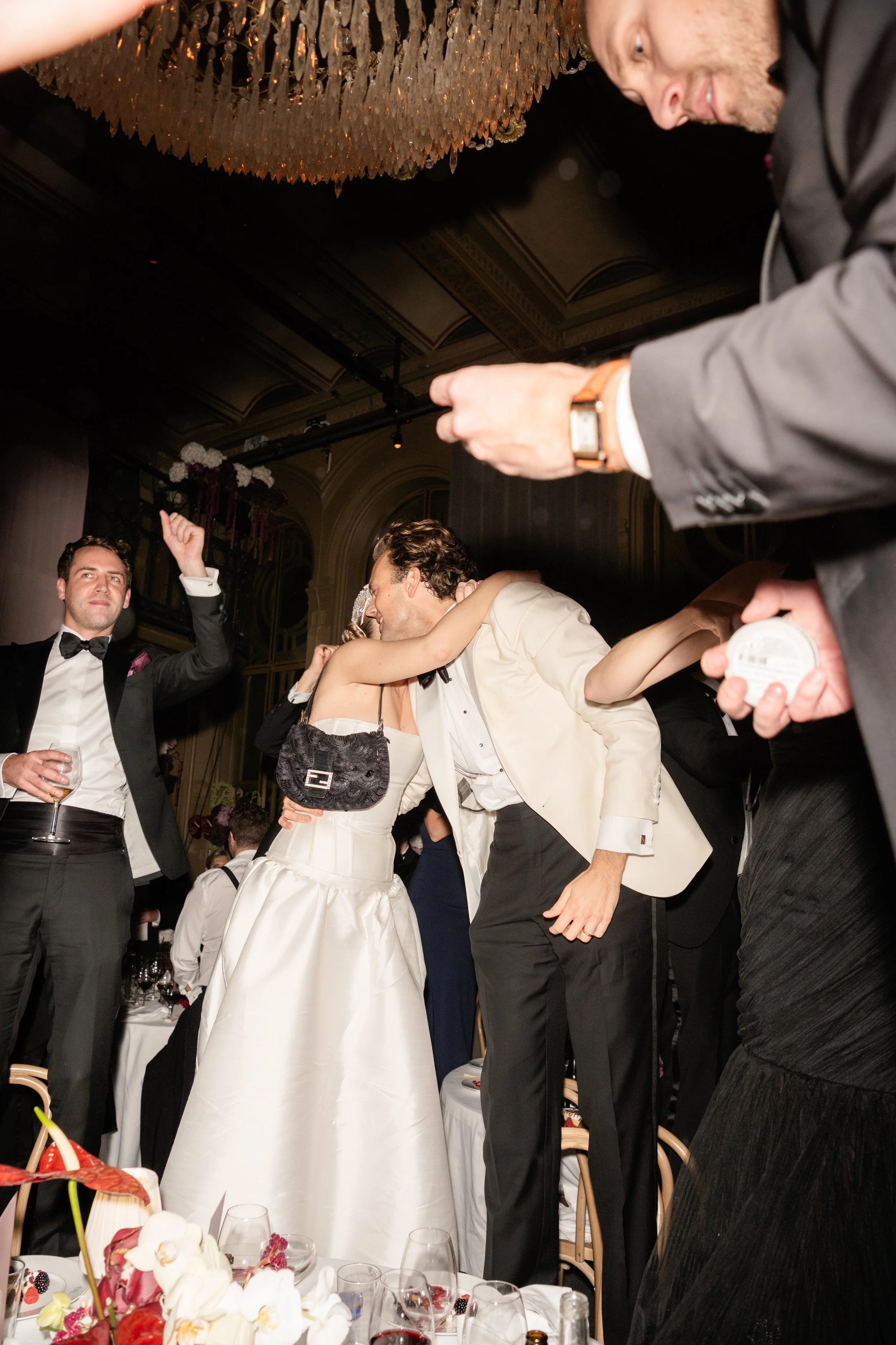 A bride and groom kissing on a table at their wedding reception, surrounded by guests in formal attire, with a man holding a microphone or recording device nearby and flowers and drinks on the table.