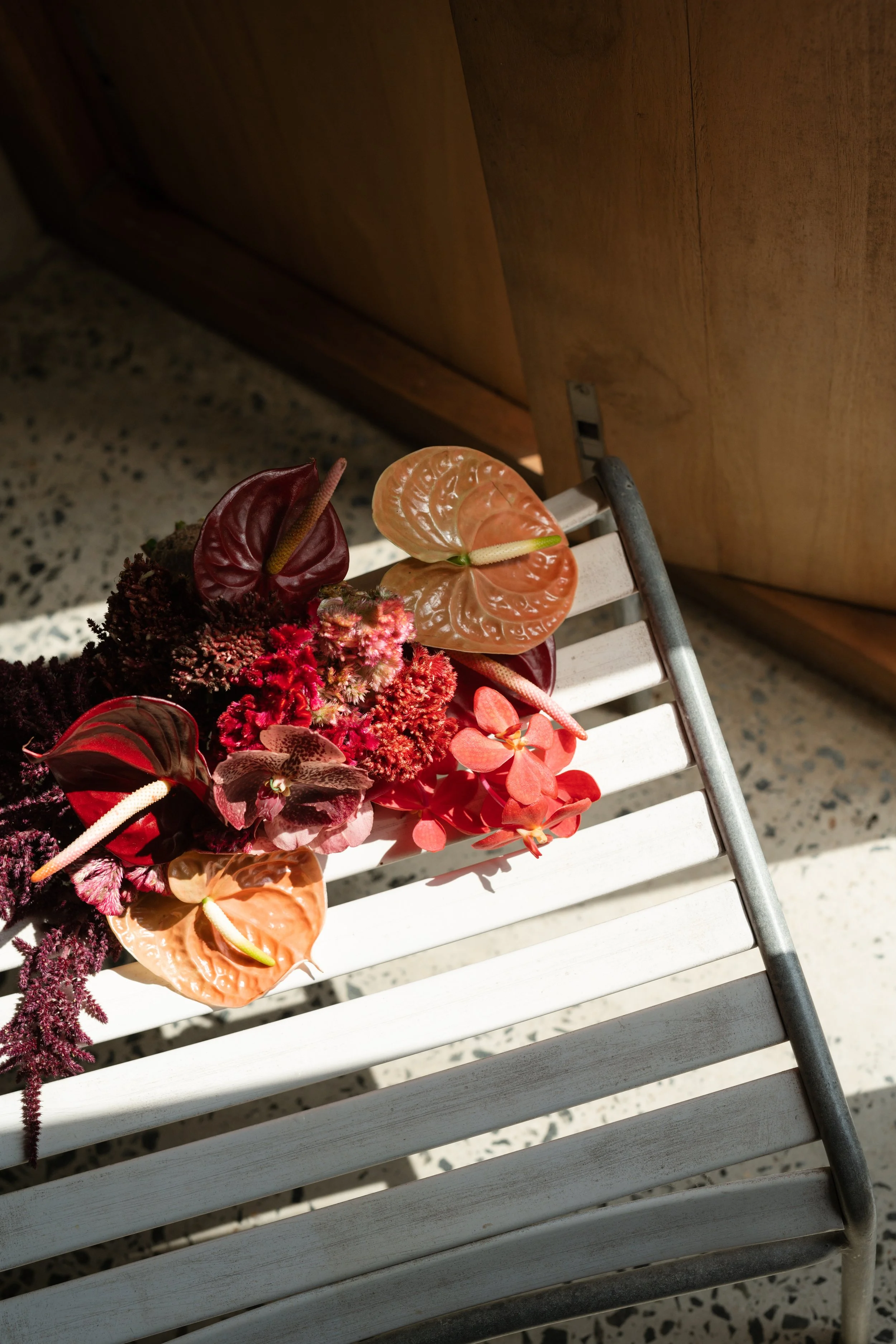 A floral arrangement with pink, red, and burgundy flowers and leaves resting on a white chair. The floor is terrazzo, and a wooden door can be seen in the background.