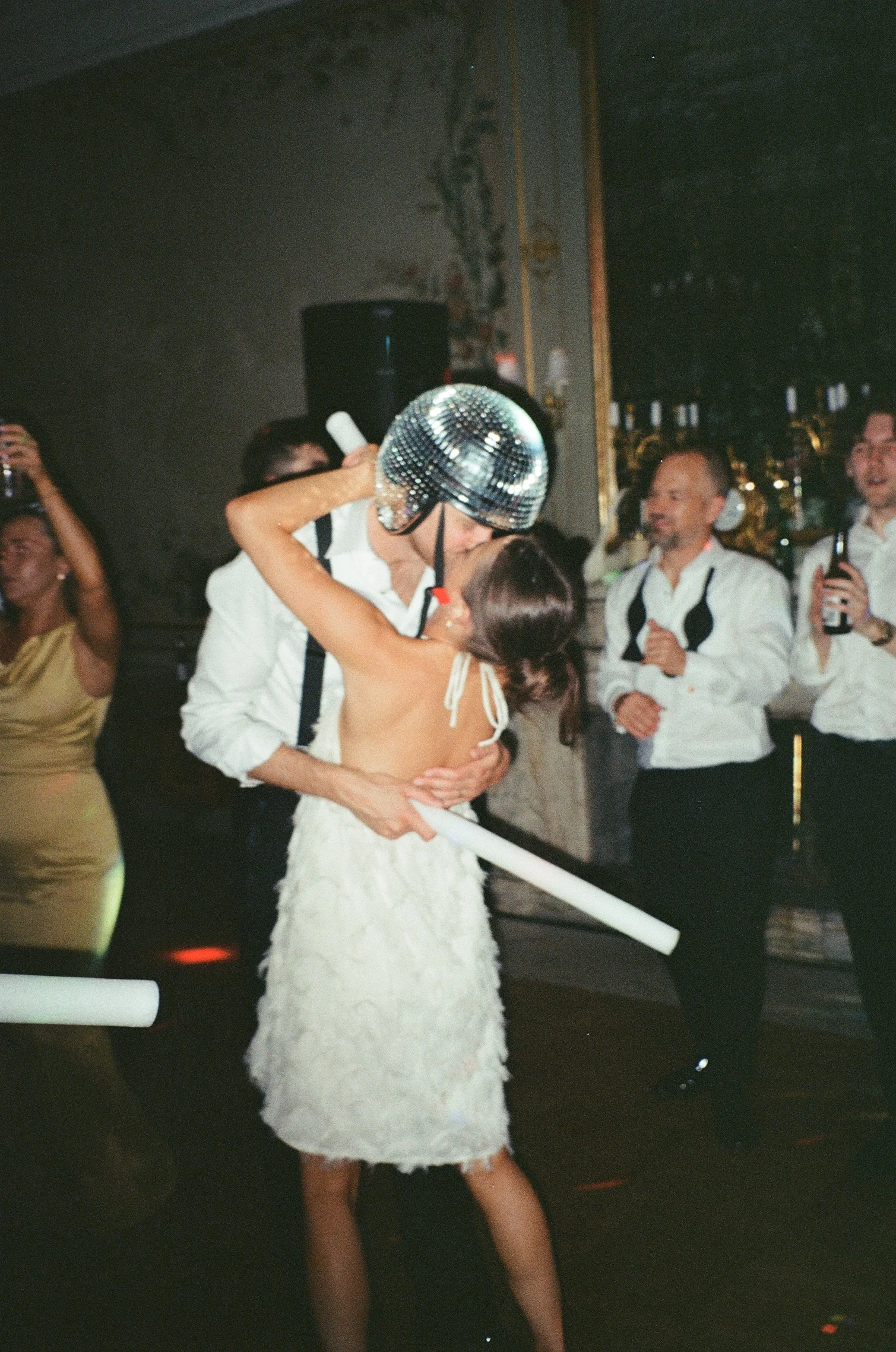People at a party, one wearing a disco ball helmet. A woman in a white dress is kissing a man. Others are dancing and taking photos.