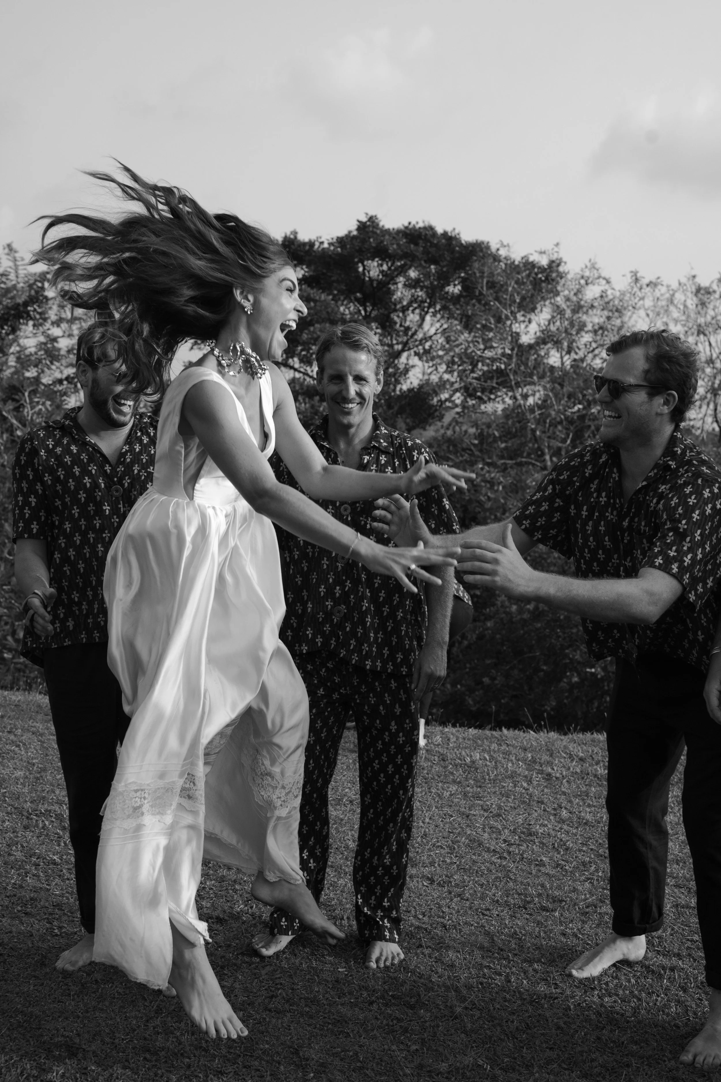 A woman in a white dress joyfully jumps with her hair flying, while four men watch and smile in an outdoor setting with trees in the background.