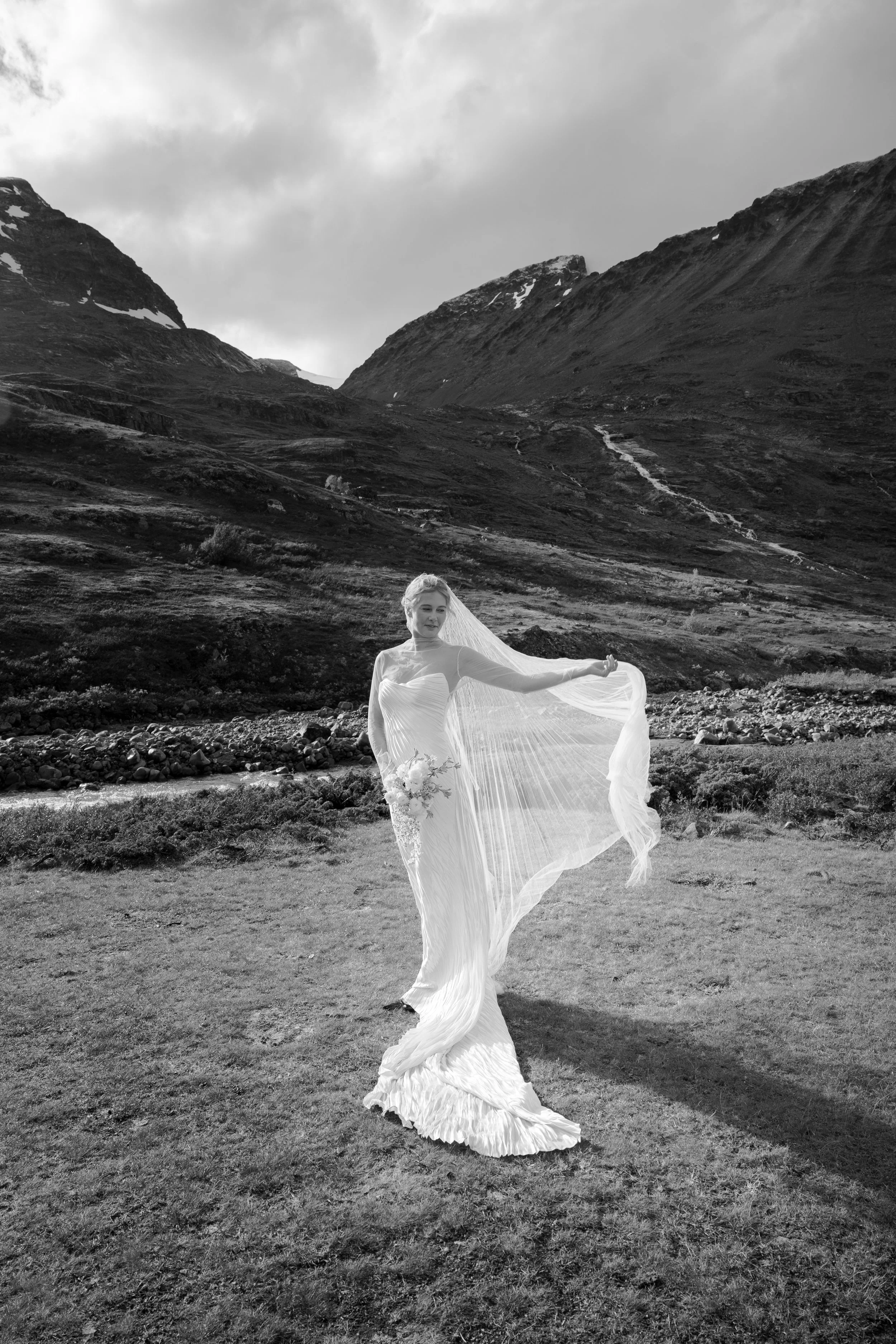 Black and white photo of a bride in a flowing wedding dress holding a bouquet, standing on grass in a mountainous landscape with cloudy sky.