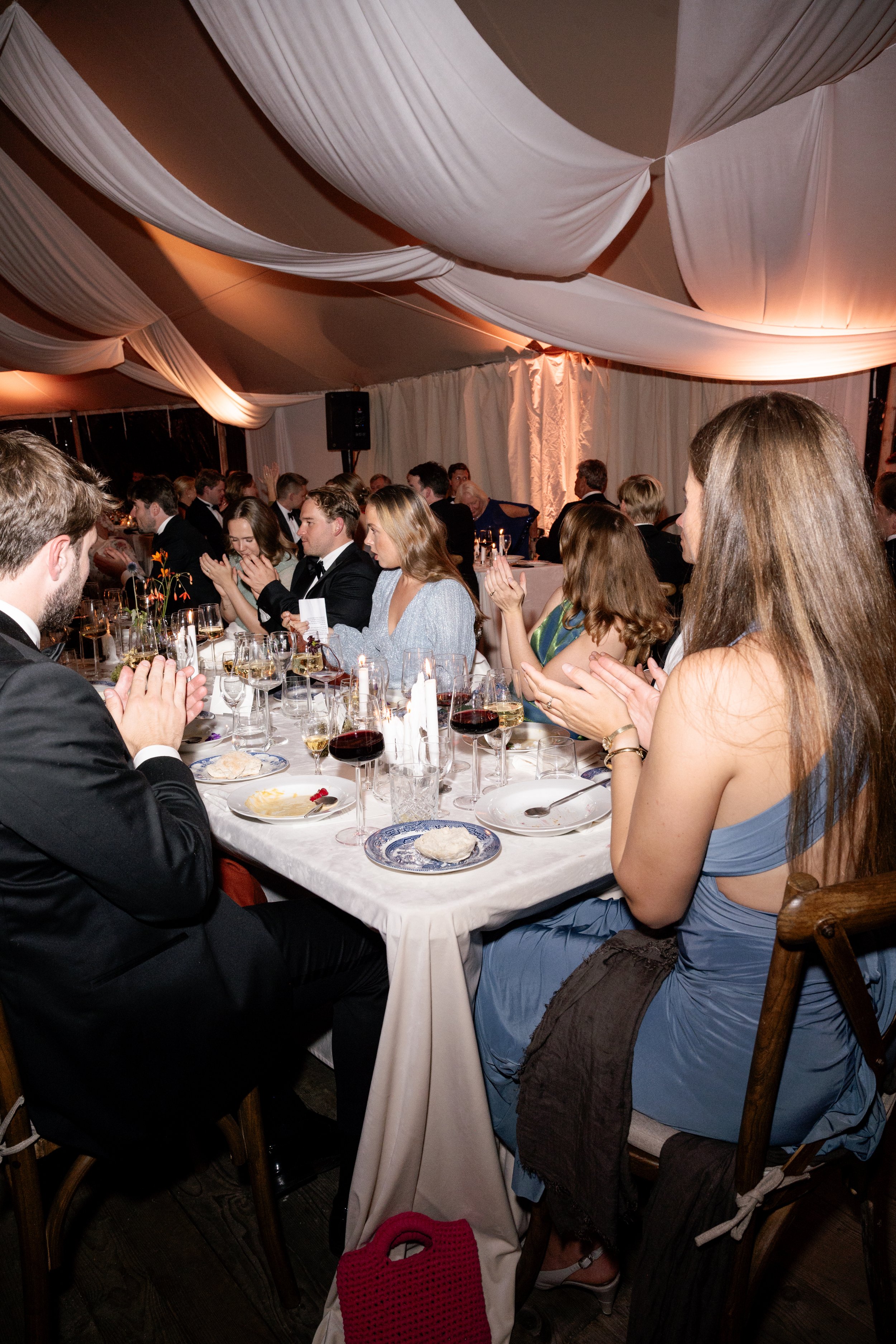 Guests at a formal dinner event sitting at a long table, clapping and enjoying their meal, under a decorated tent canopy.