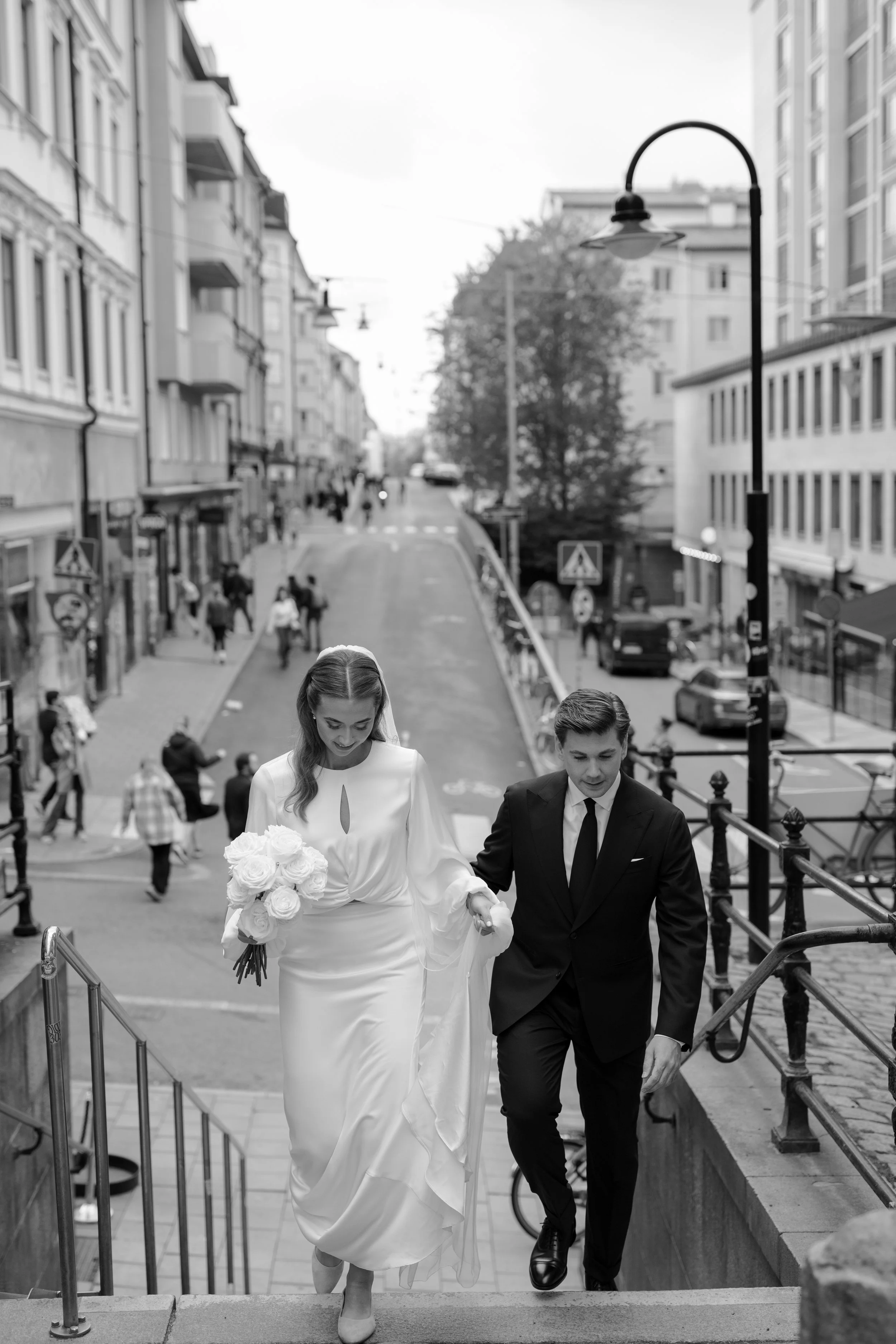 A bride and groom walking up stairs on a city street, with people and cars in the background, in black and white.