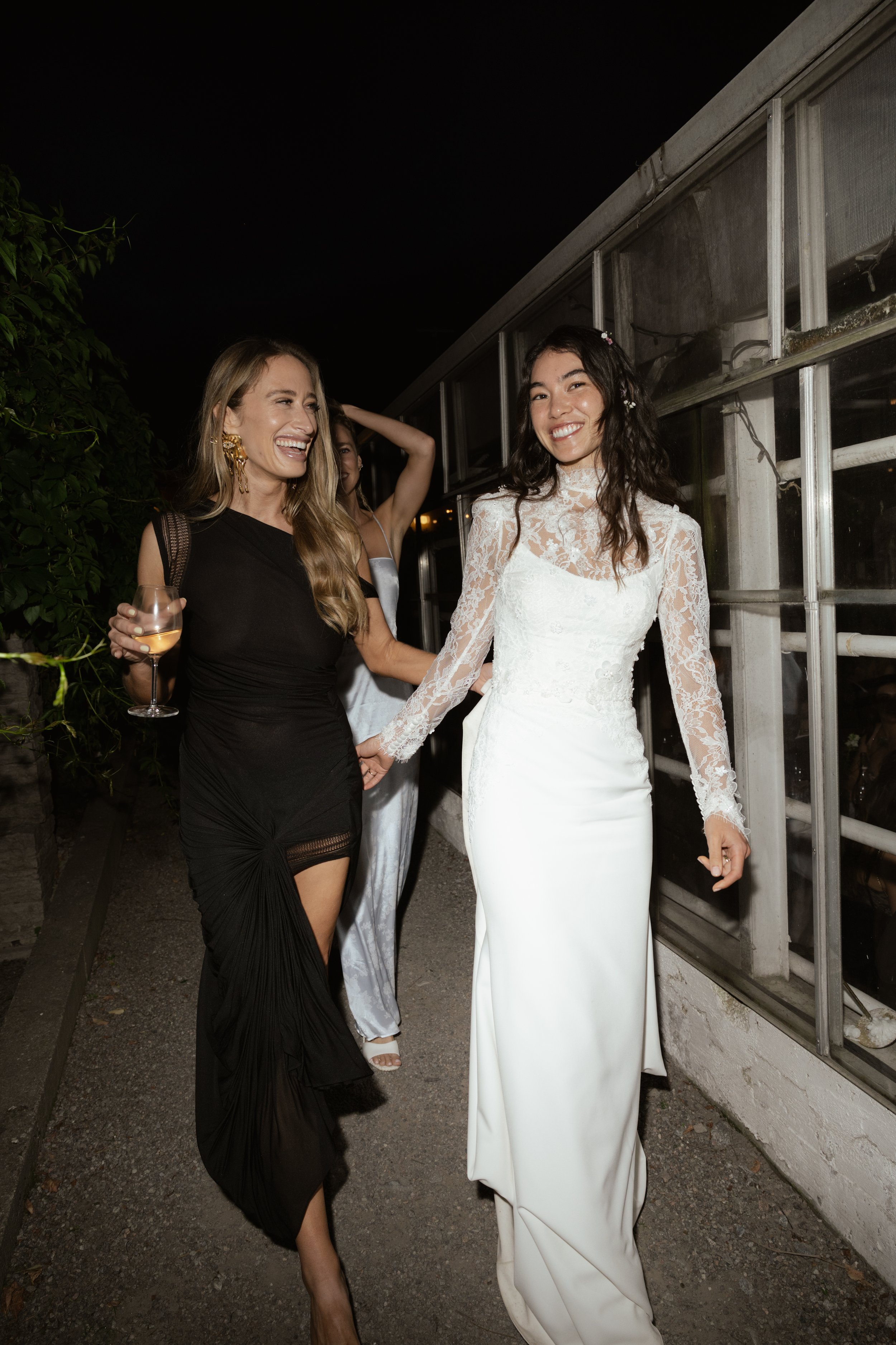 Two women holding hands and smiling, one in a white wedding dress with lace details, the other in a black dress, at night outdoors.