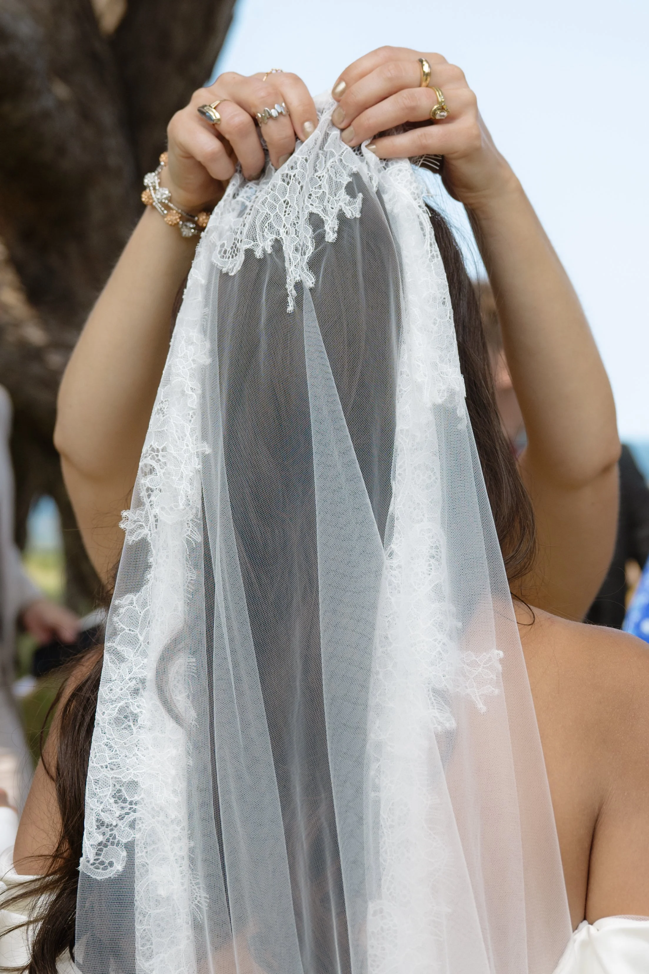 A woman placing a white lace veil over her head, adjusting it with both hands, wearing multiple rings and a bracelet, with a tree trunk and a blurred outdoor background.