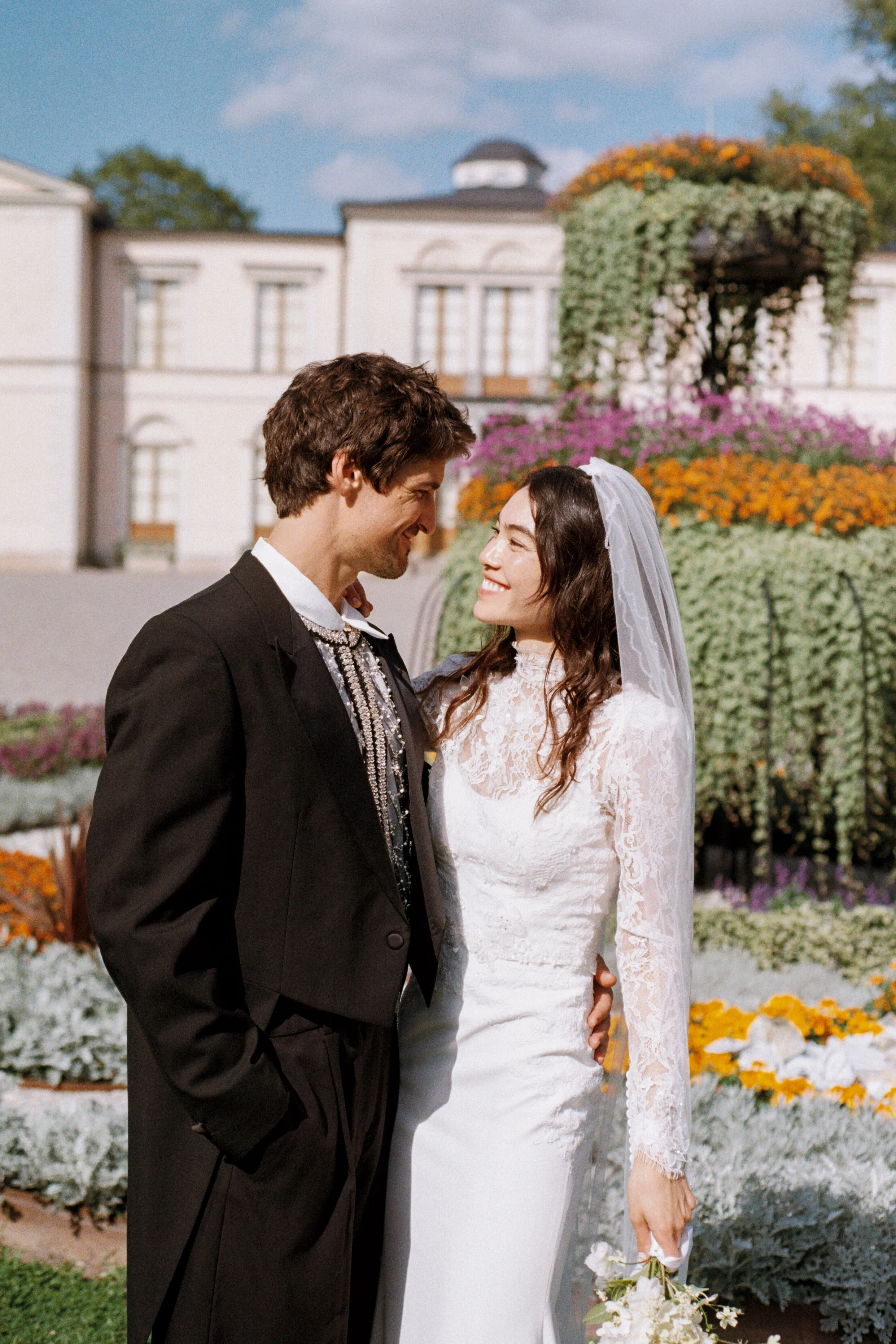A bride and groom smiling at each other during their wedding in a garden with colorful flowers and a white building in the background.