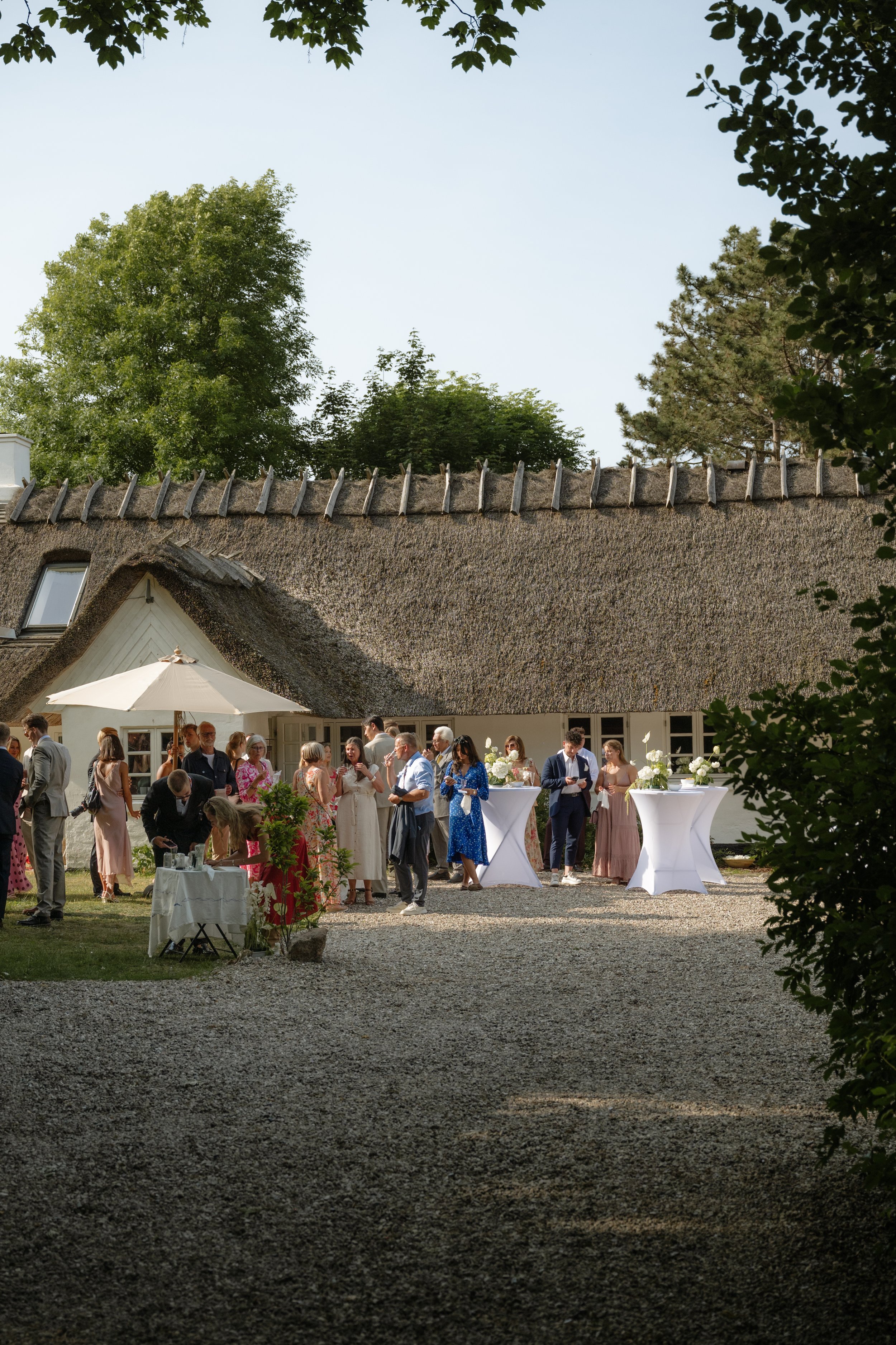 People gathered outdoors for a celebration near a thatched-roof building, some standing around high tables with white tablecloths and floral arrangements, under a sunny sky.