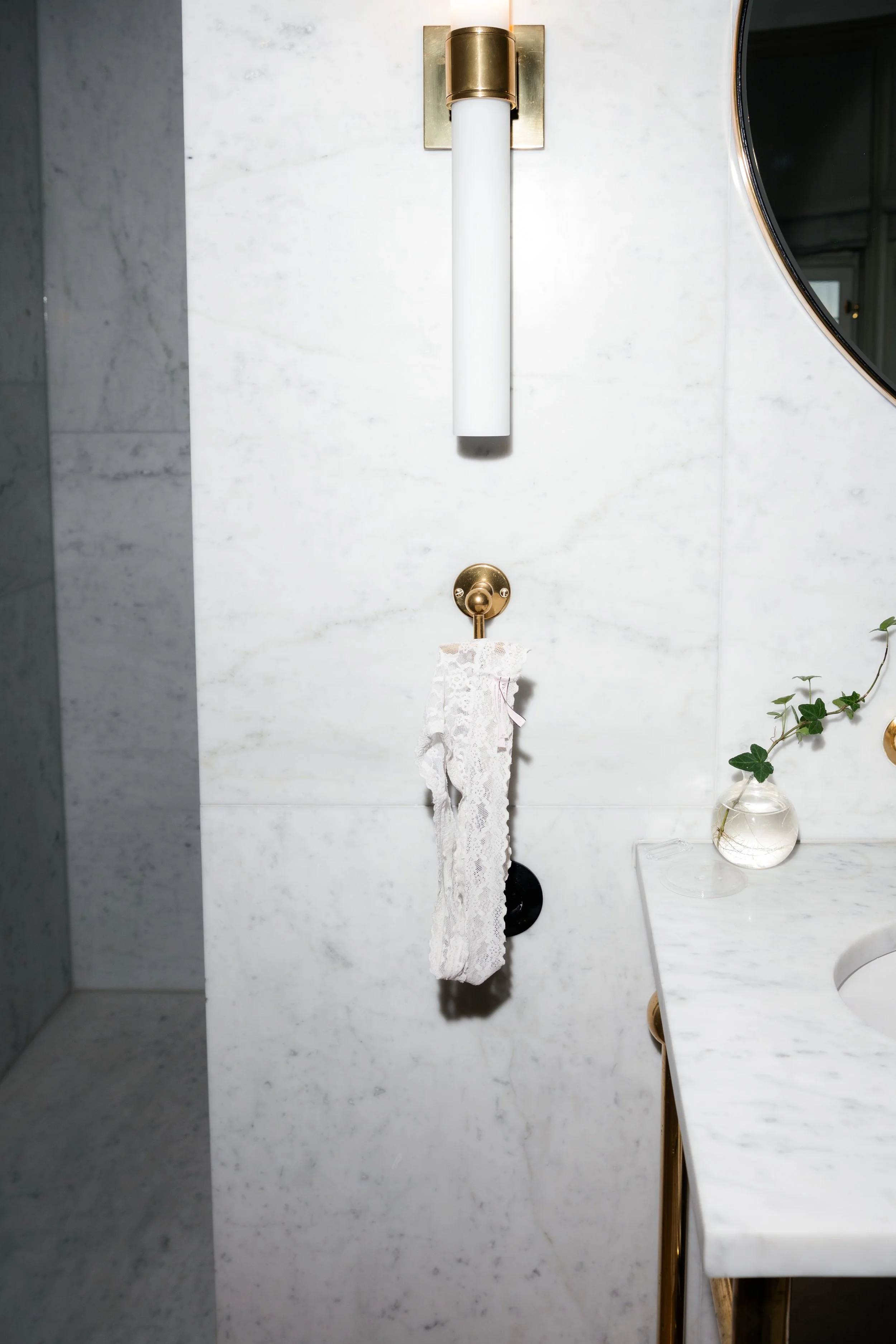 Modern bathroom wall with a gold fixtures, a white towel hanging from a hook, a small glass vase with green ivy on a marble countertop, and a round mirror.