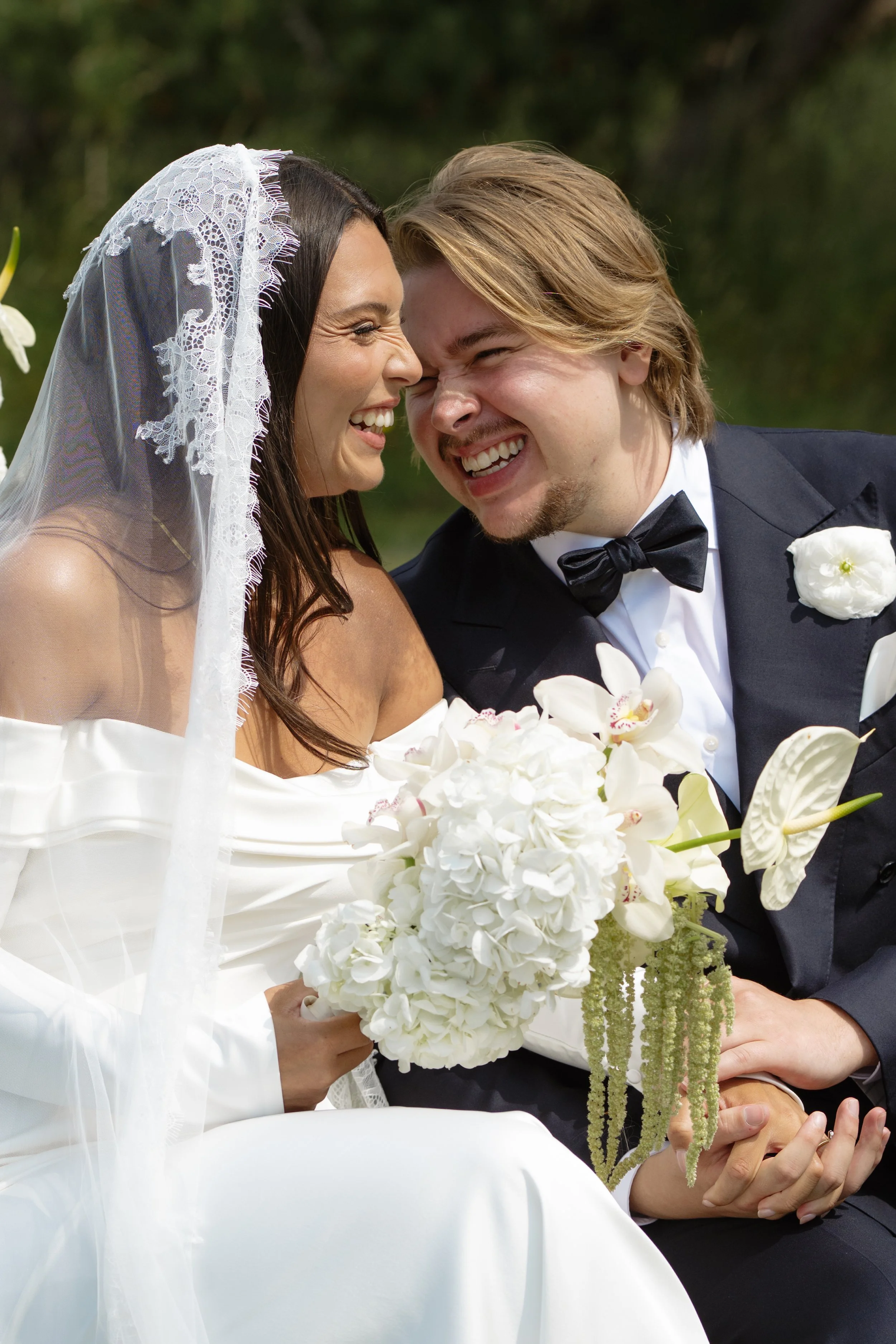 A bride and groom laughing closely during their wedding, with the bride holding a bouquet of white flowers and the groom wearing a tuxedo with a bow tie.