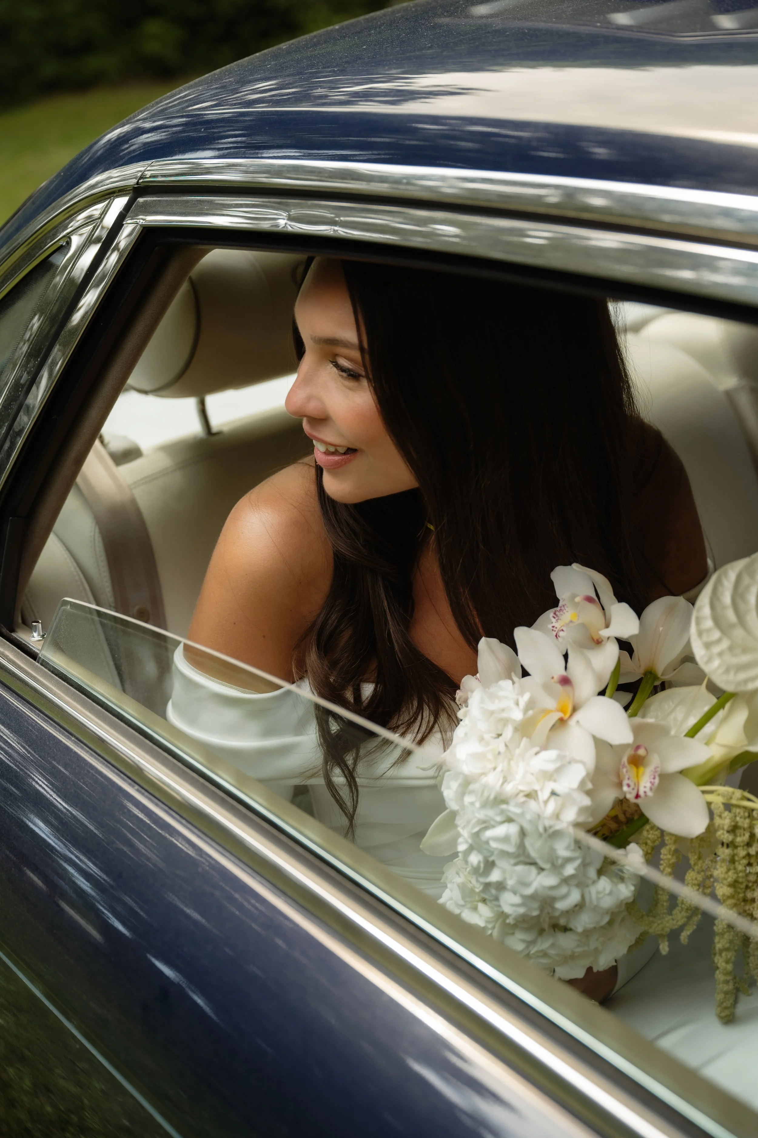 A woman with dark hair smiling and sitting in a vintage car, holding a bouquet of white flowers like orchids and hydrangeas, on a green outdoor setting.