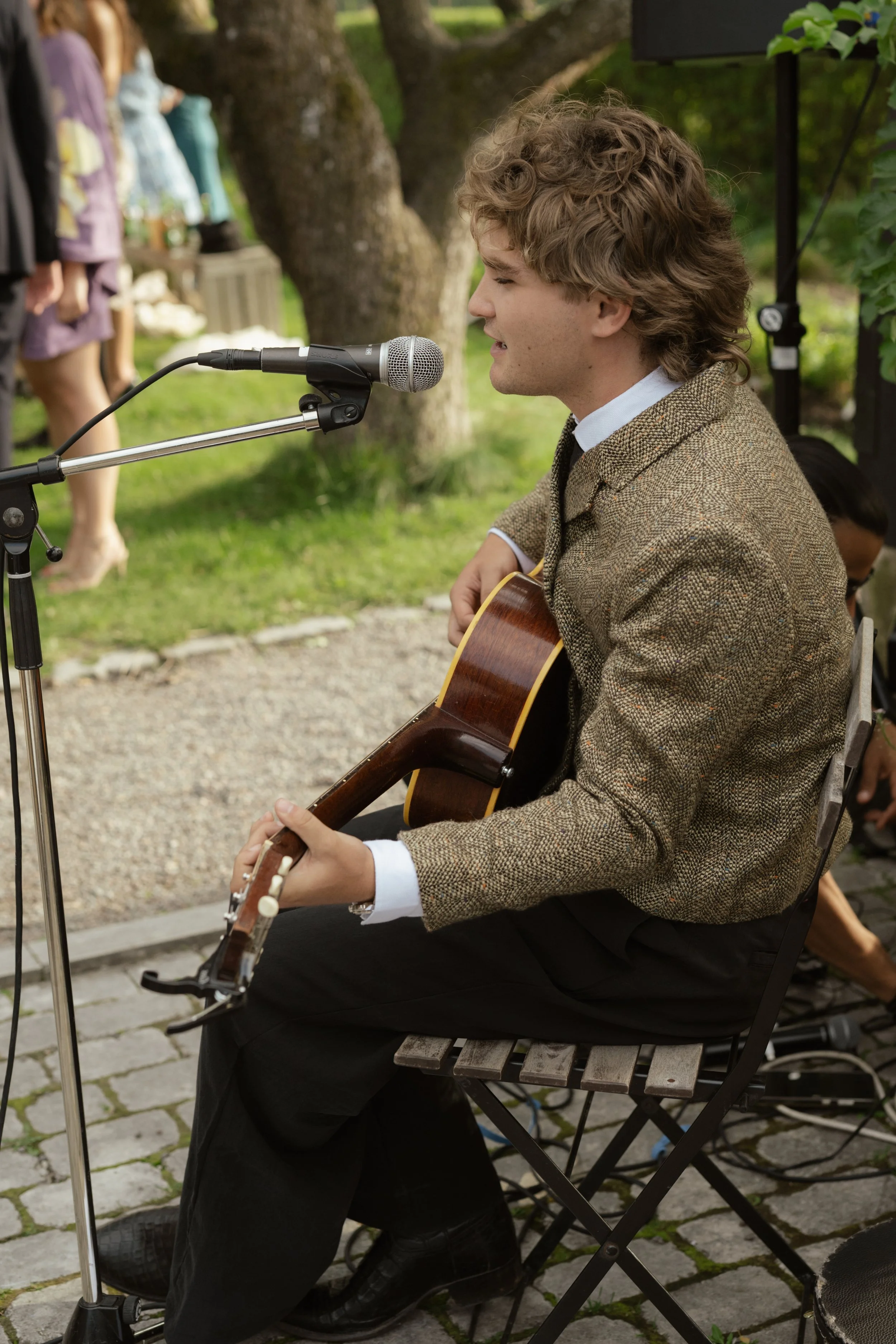 A young man with curly hair playing an acoustic guitar and singing into a microphone while sitting on a folding chair outdoors during the daytime.