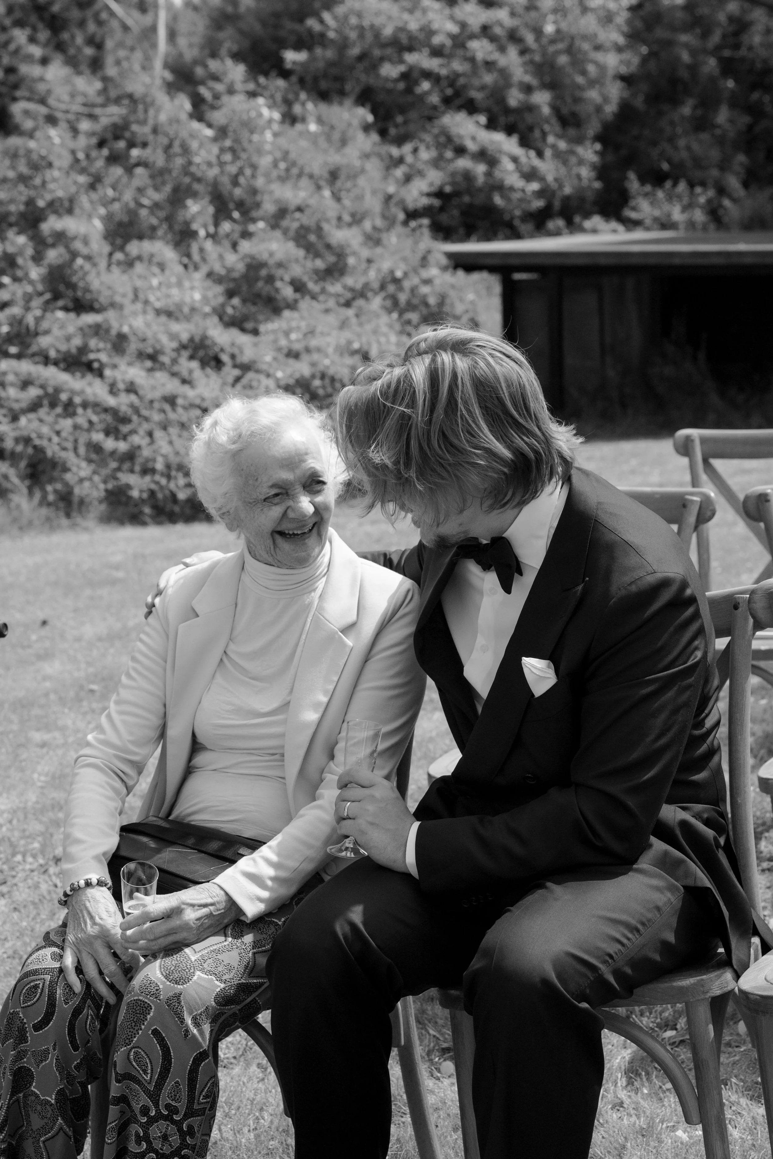 An elderly woman and a young man in a tuxedo sit outdoors, smiling and laughing together, holding glasses, with trees and a building in the background.