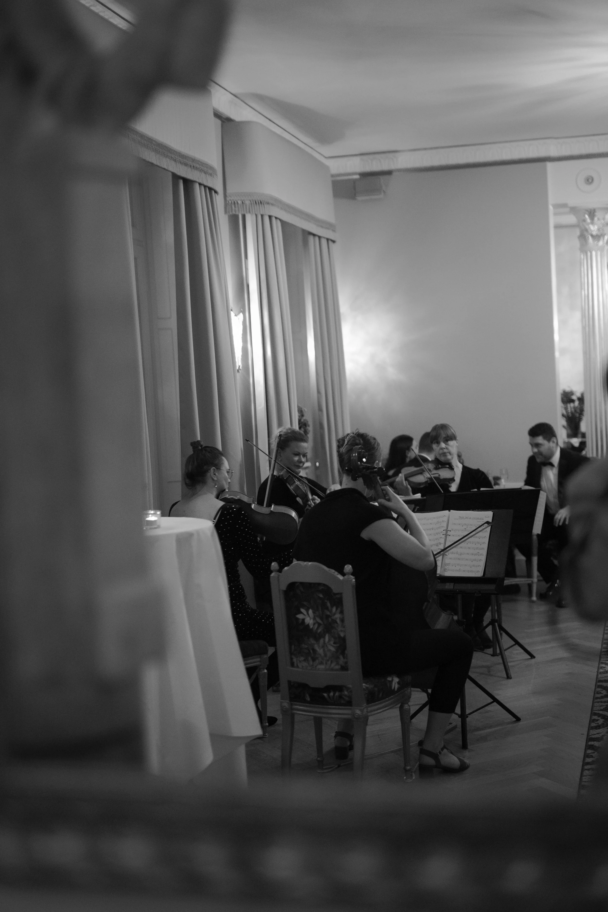 A black and white photo of a classical music ensemble performing indoors, with chandeliers, curtains, and ornate decorations visible.