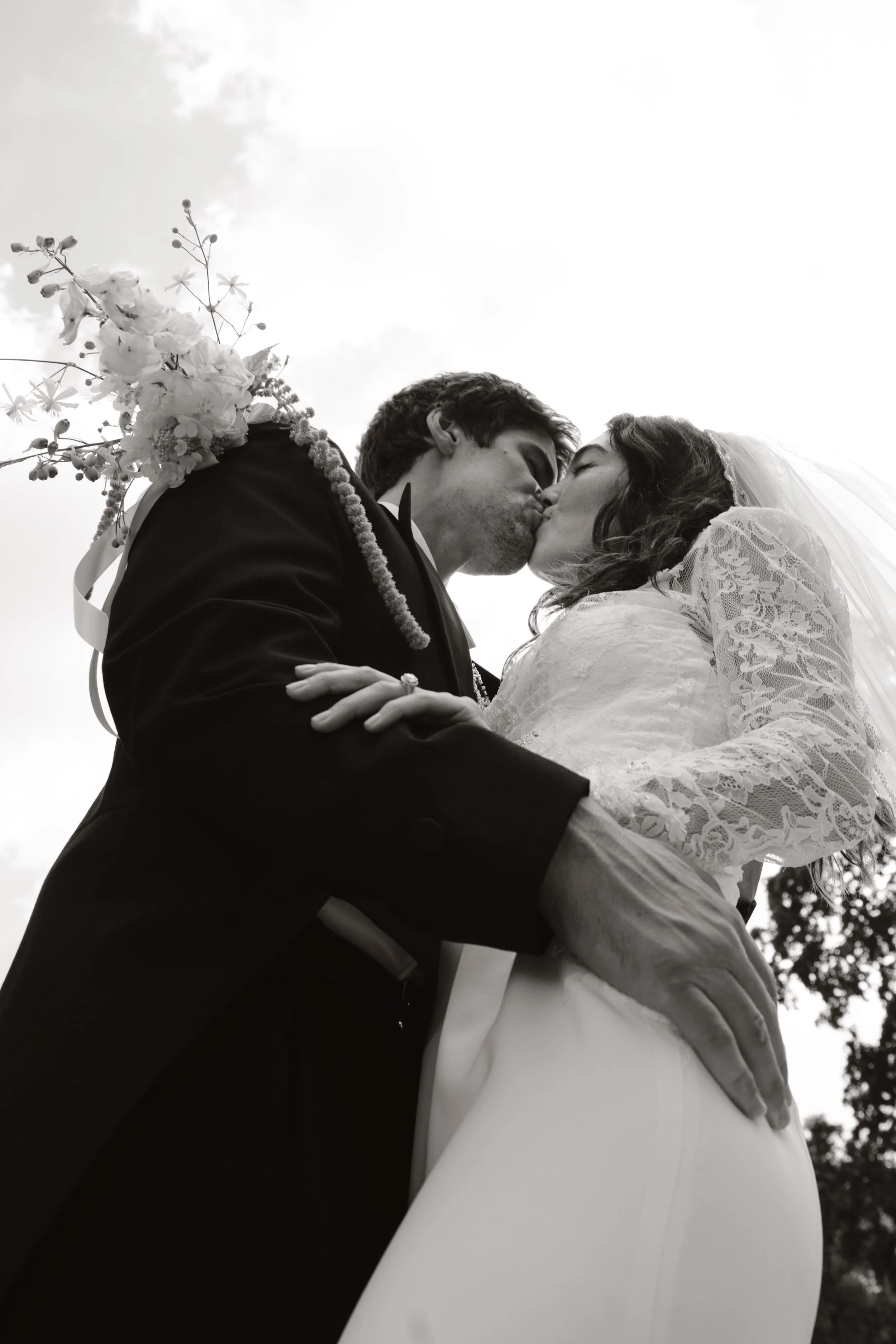 A bride and groom kissing outdoors, with the photo taken from below, showing the bride wearing a lace wedding dress and veil, and the groom in a dark suit. The bride holds a floral bouquet over her shoulder.