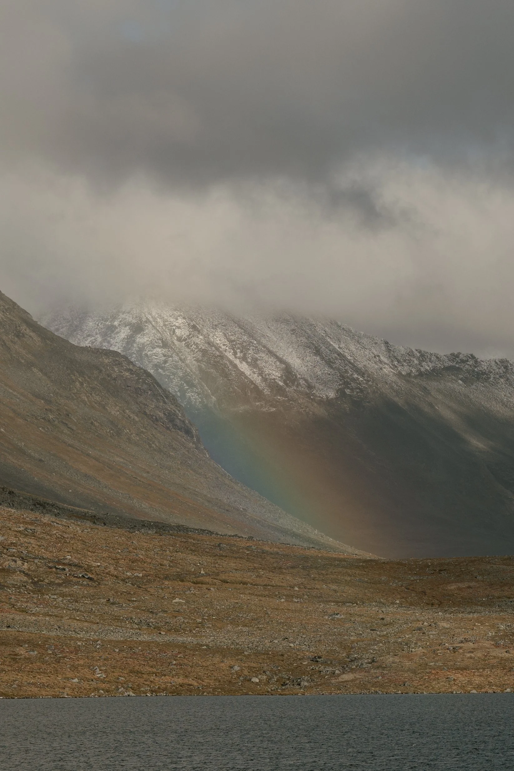 Snow-capped mountains with a rainbow visible on their slope under gray clouds, next to a dark body of water.