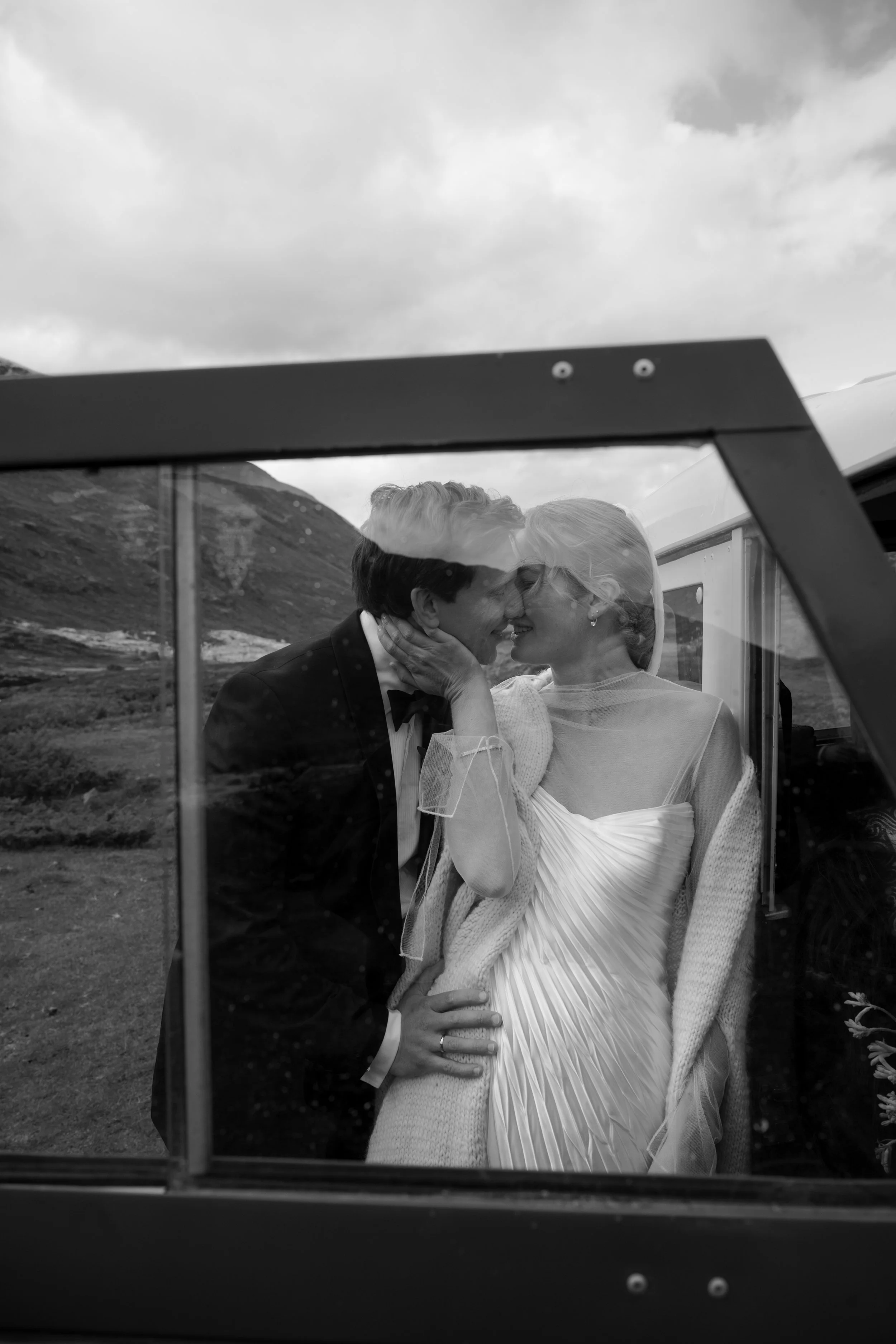 A black and white photo of a couple, likely newlyweds, sharing an intimate moment inside a window reflection with mountains in the background. The man is in a tuxedo and the woman in a wedding dress, leaning close with their faces touching.