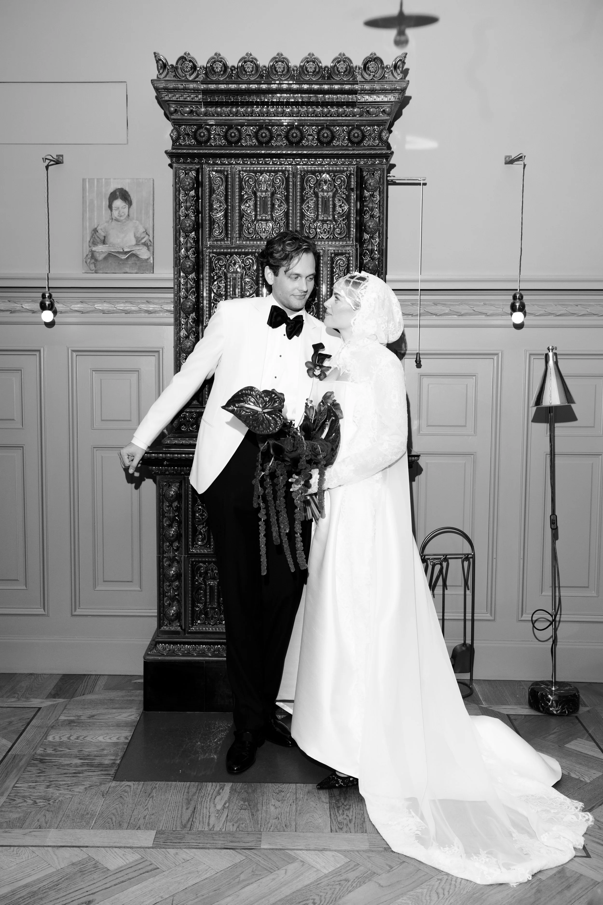 A black-and-white photo of a bride and groom standing close together in an elegant room. The groom is dressed in a tuxedo, and the bride is in a long wedding gown holding a large bouquet. They are looking into each other's eyes.