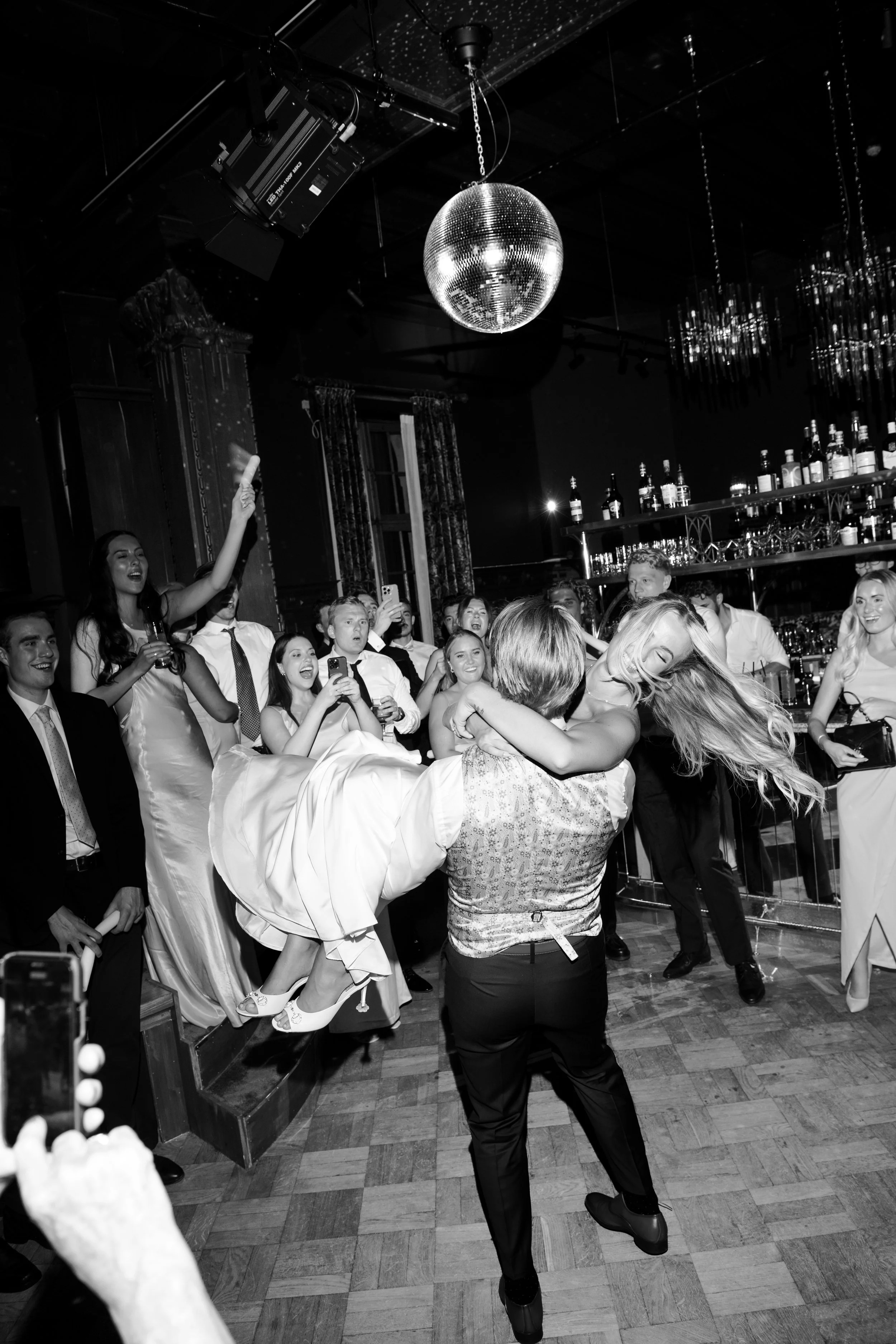 People dancing and celebrating at a party in a tuxedo and formal dresses with a disco ball overhead.