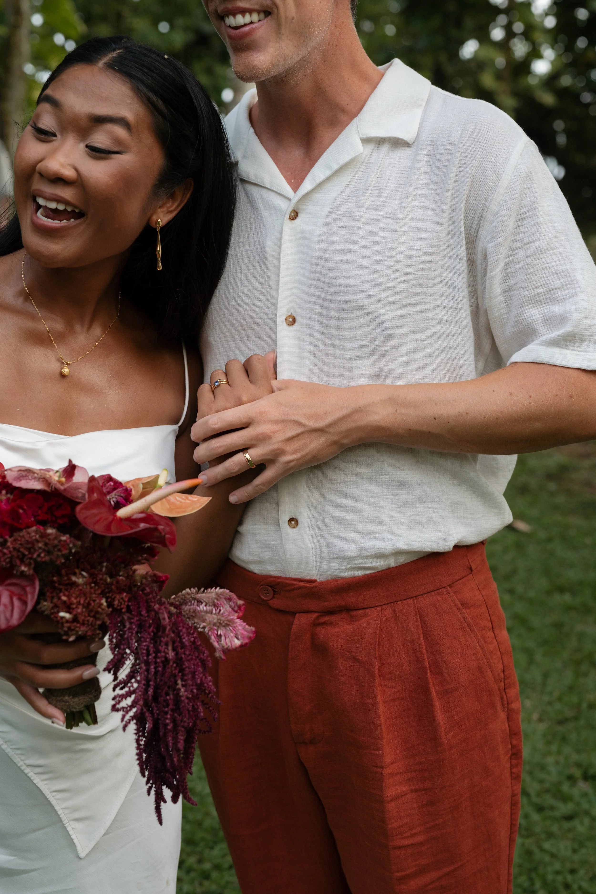 A woman in a white dress holding a colorful flower bouquet, standing next to a man in a white shirt and red pants, holding hands outdoors.