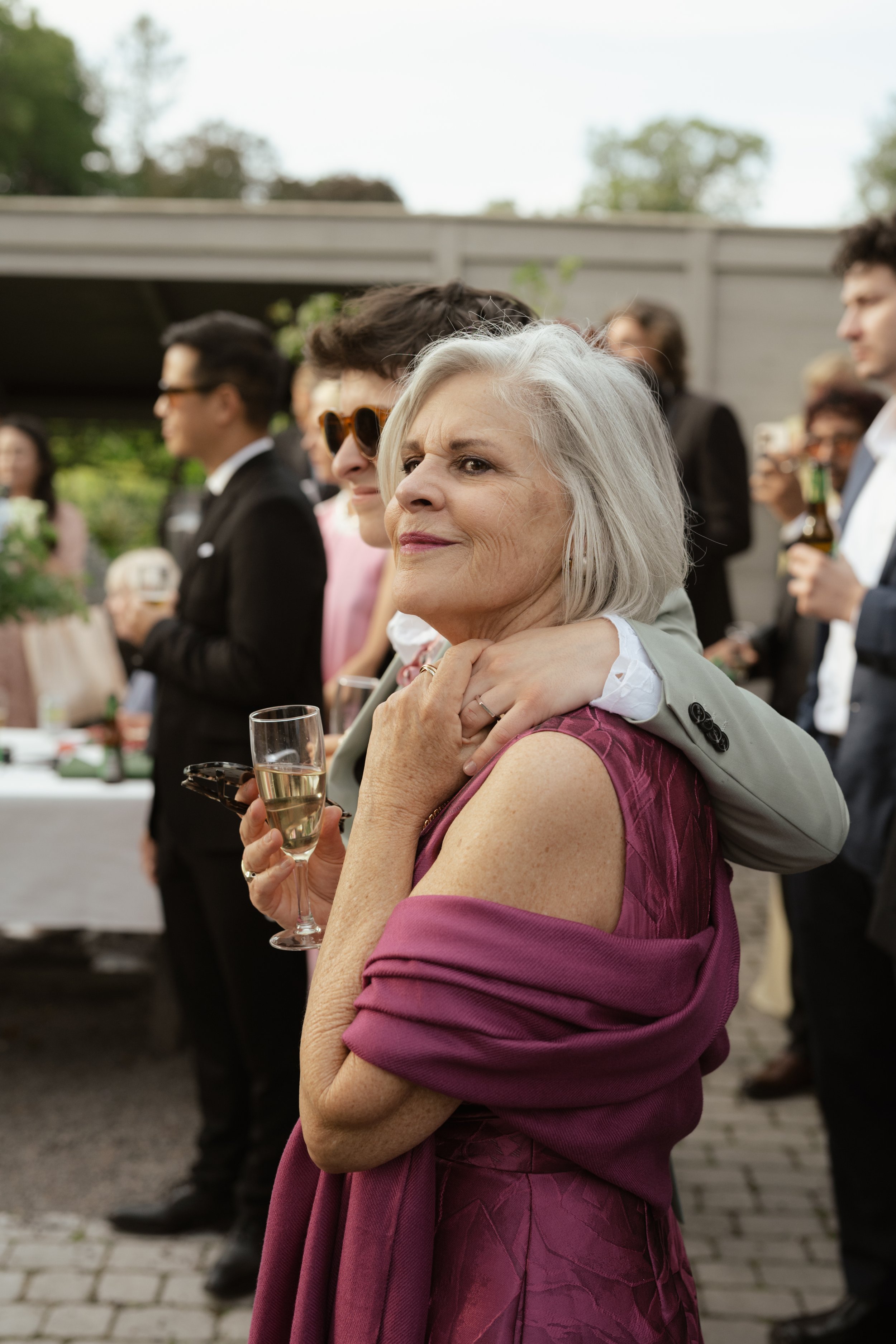 An elderly woman with gray hair holding a glass of champagne at an outdoor gathering, surrounded by people dressed formally.
