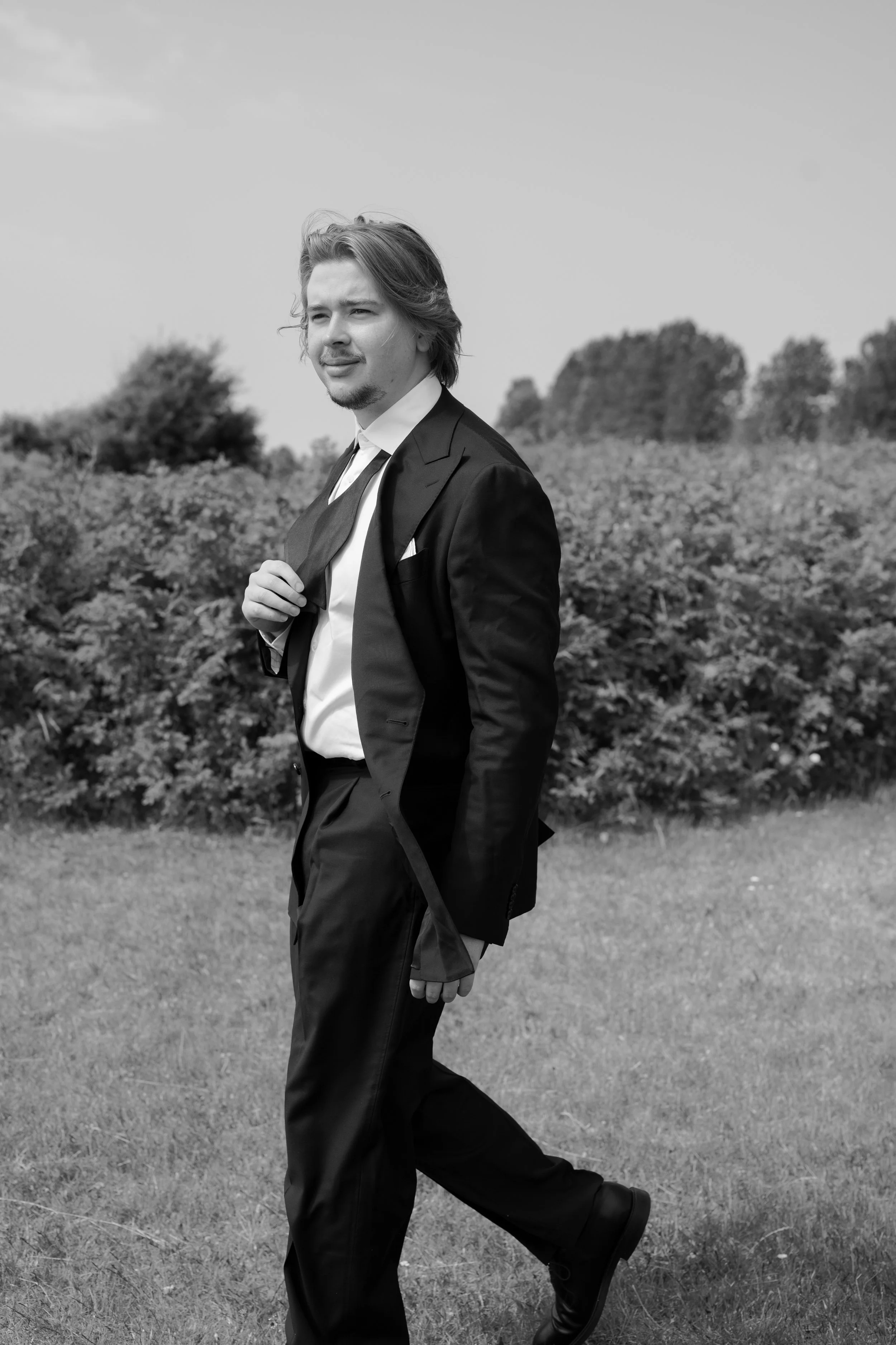 A young man in a suit walking outdoors on a grassy field with bushes and trees in the background, holding his jacket over his shoulder.