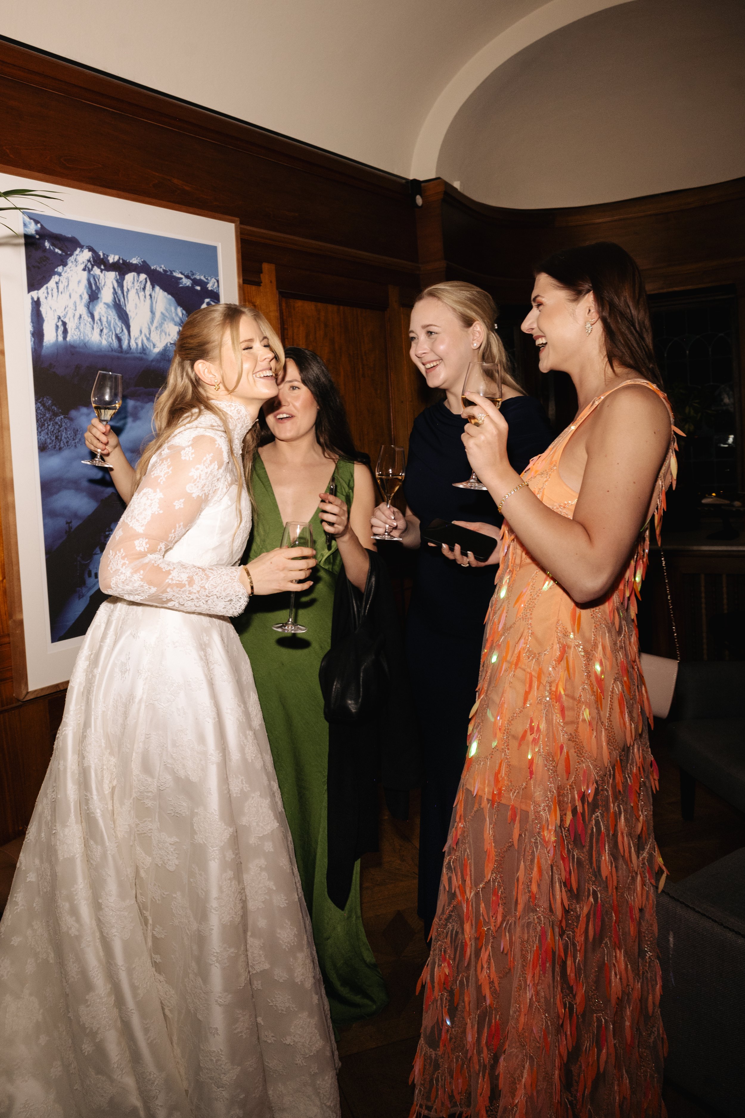 A group of four women in elegant dresses celebrating at a social gathering, holding glasses of champagne and laughing.