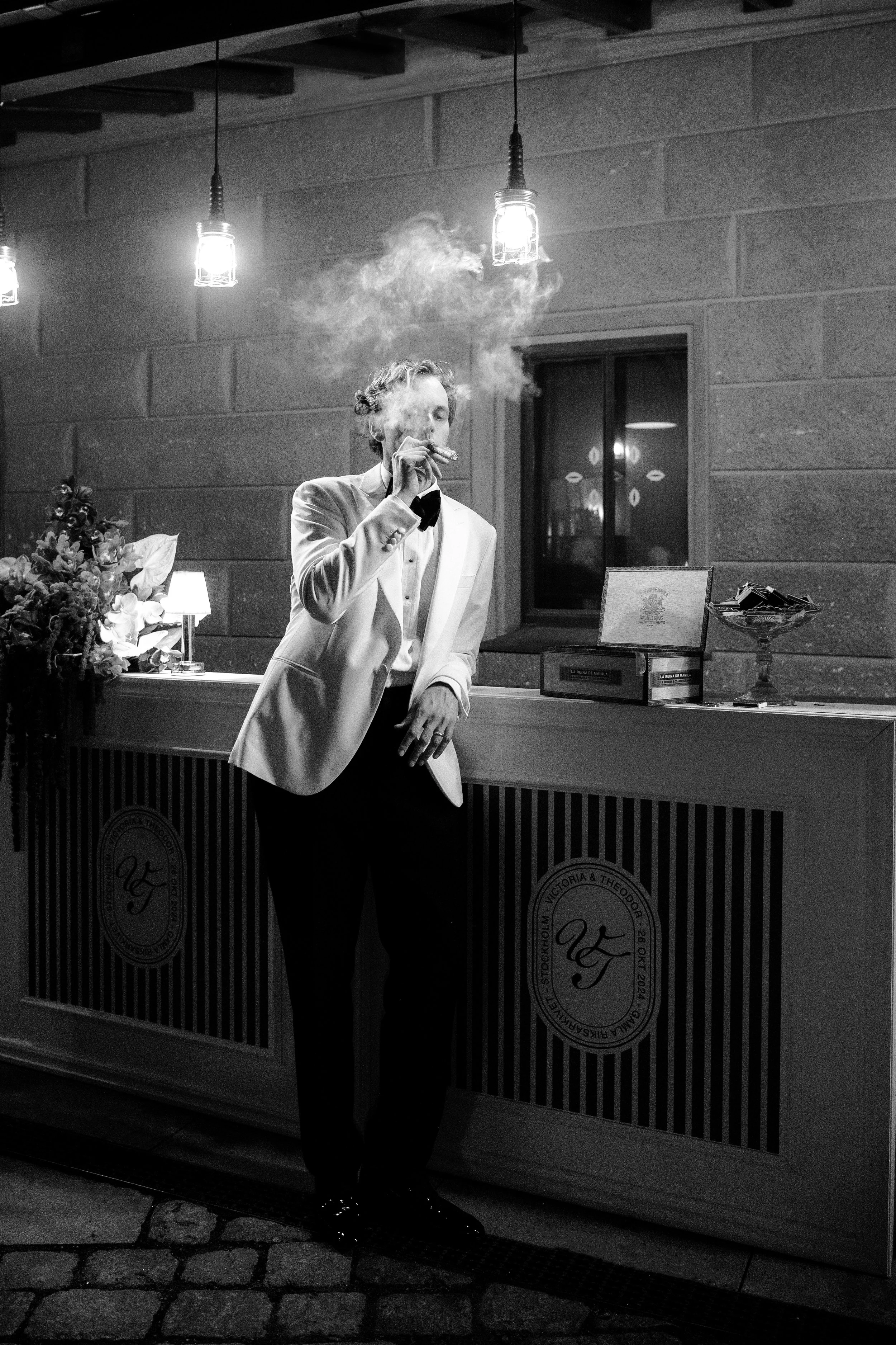 A man in a tuxedo and white blazer smokes a cigar indoors at a reception area, with bright hanging lights and a flower arrangement visible.
