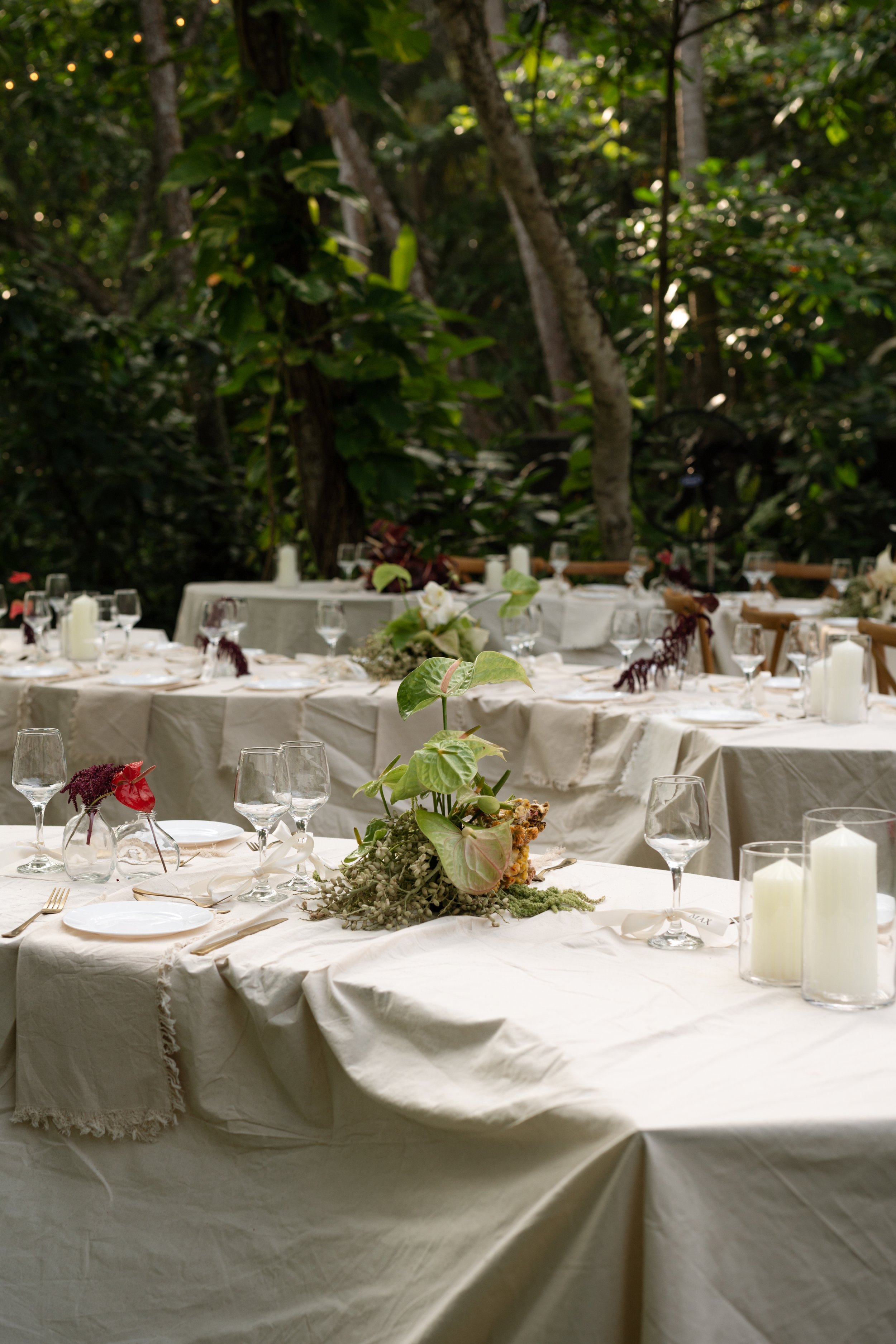 Elegant outdoor dining reception with long tables decorated with white tablecloths, glassware, candles, and floral centerpieces set in a lush green forest.