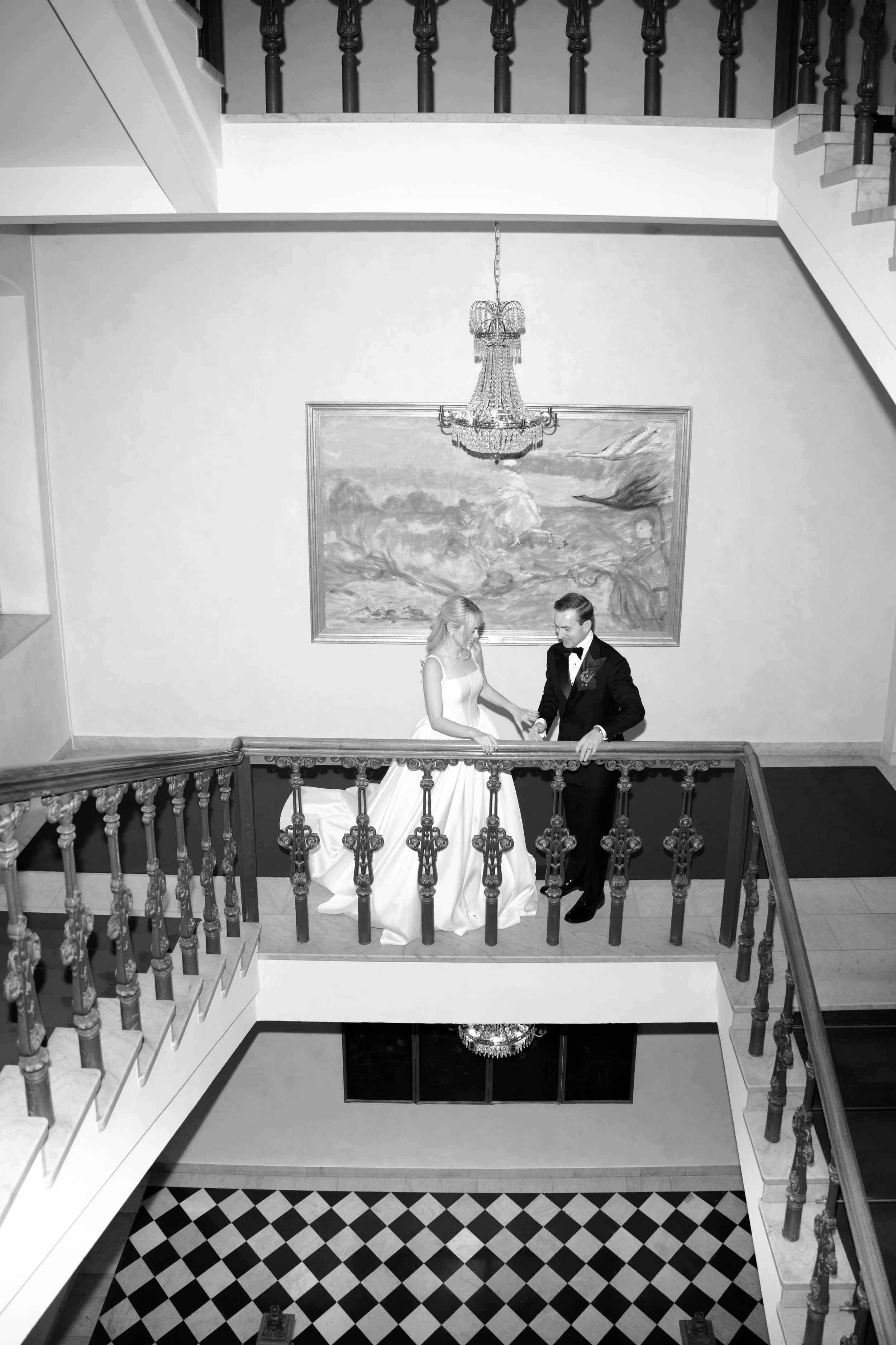 Bride and groom holding hands on a staircase landing, with a chandelier and a large framed painting in the background, in black and white.