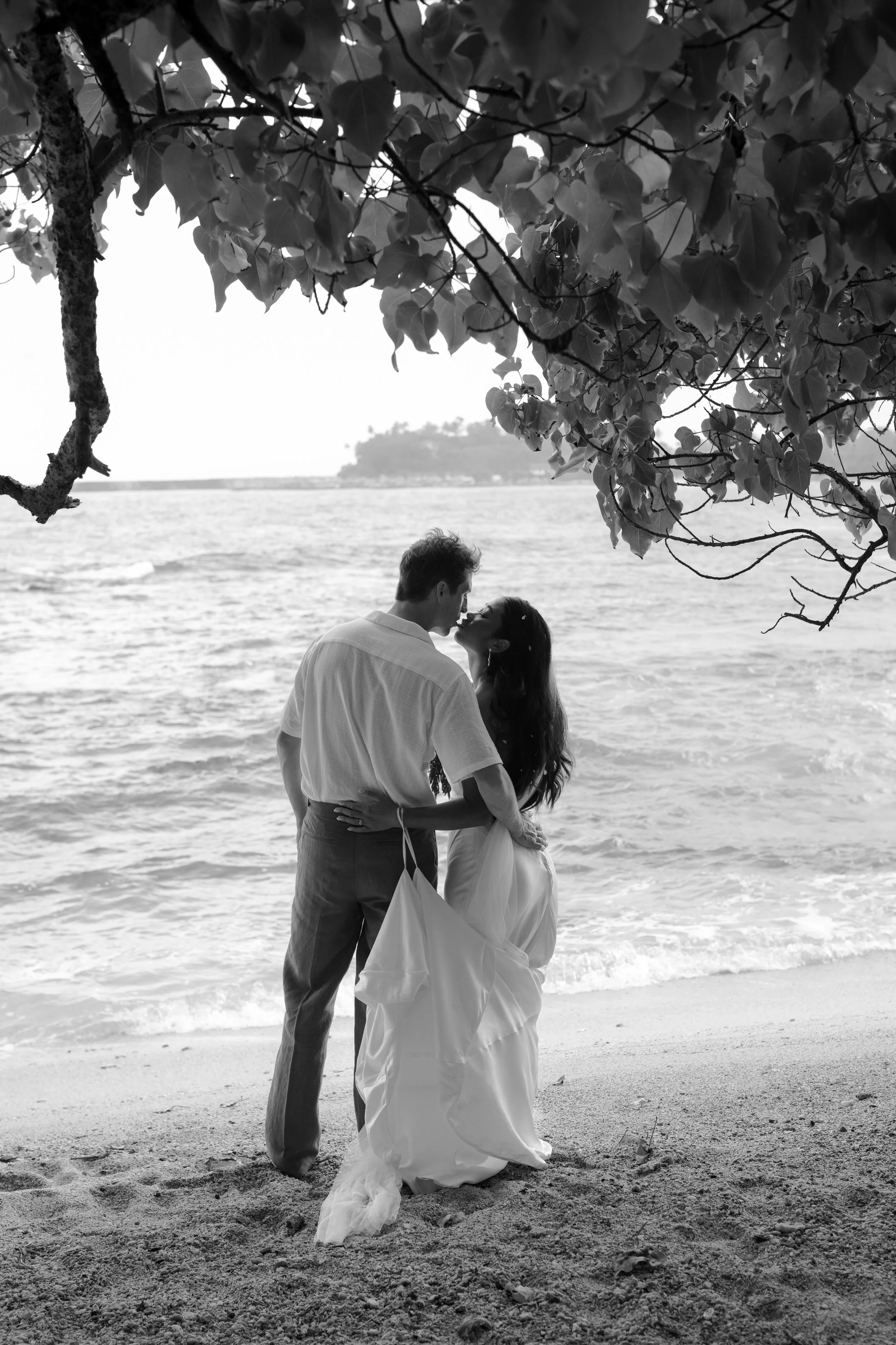 A black-and-white photo of a couple kissing on a beach, under a large tree with leaves hanging down, near the water with some land in the background.