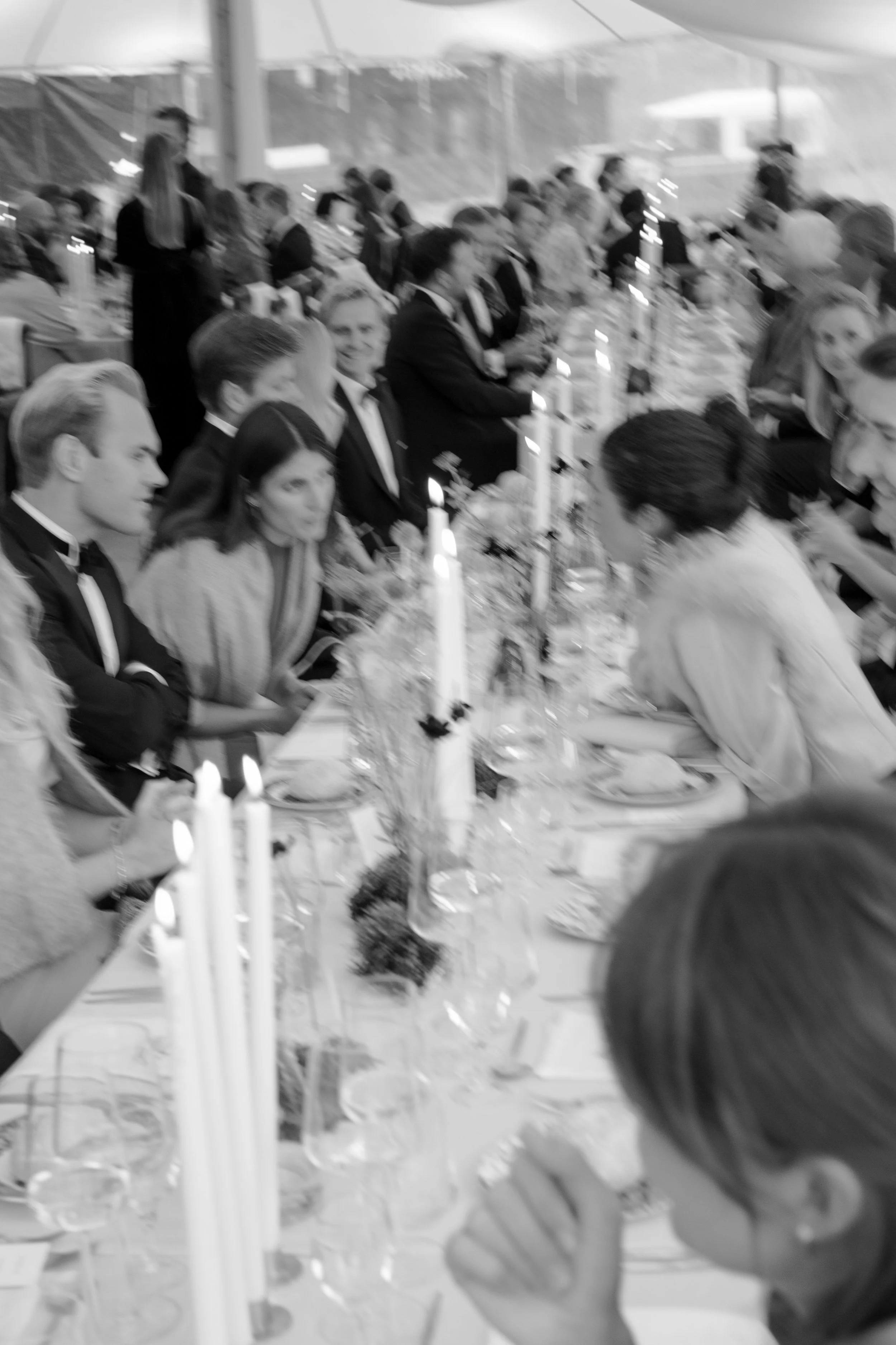 People dressed in formal attire, seated at a long banquet table with candles and floral centerpieces, in a tented outdoor setting.