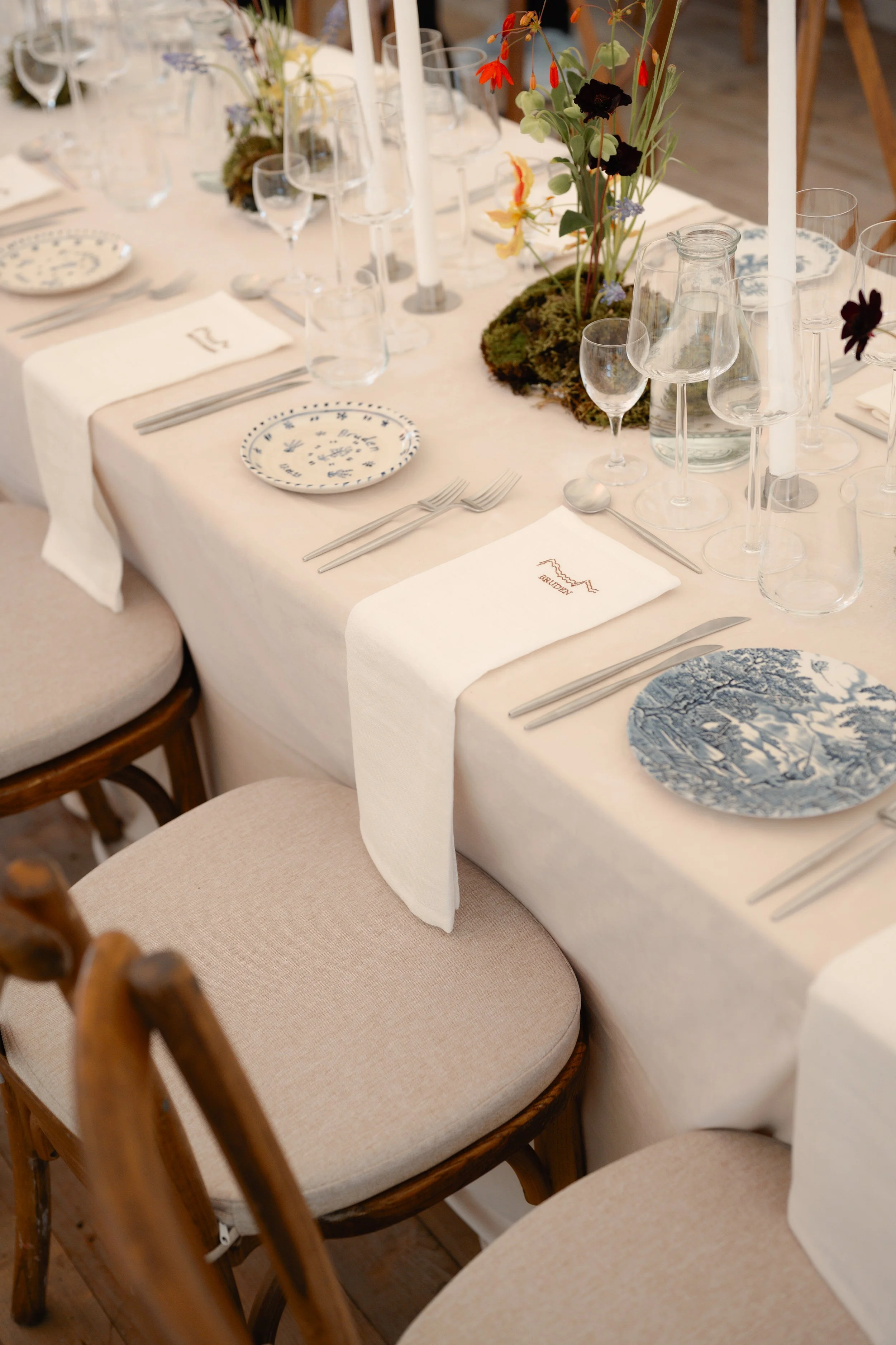 Elegant banquet table set for a special occasion with white tablecloth, white napkins, and various glassware, plates, and cutlery, with a floral centerpiece featuring red, orange, dark purple, and blue flowers, and tall white candles.