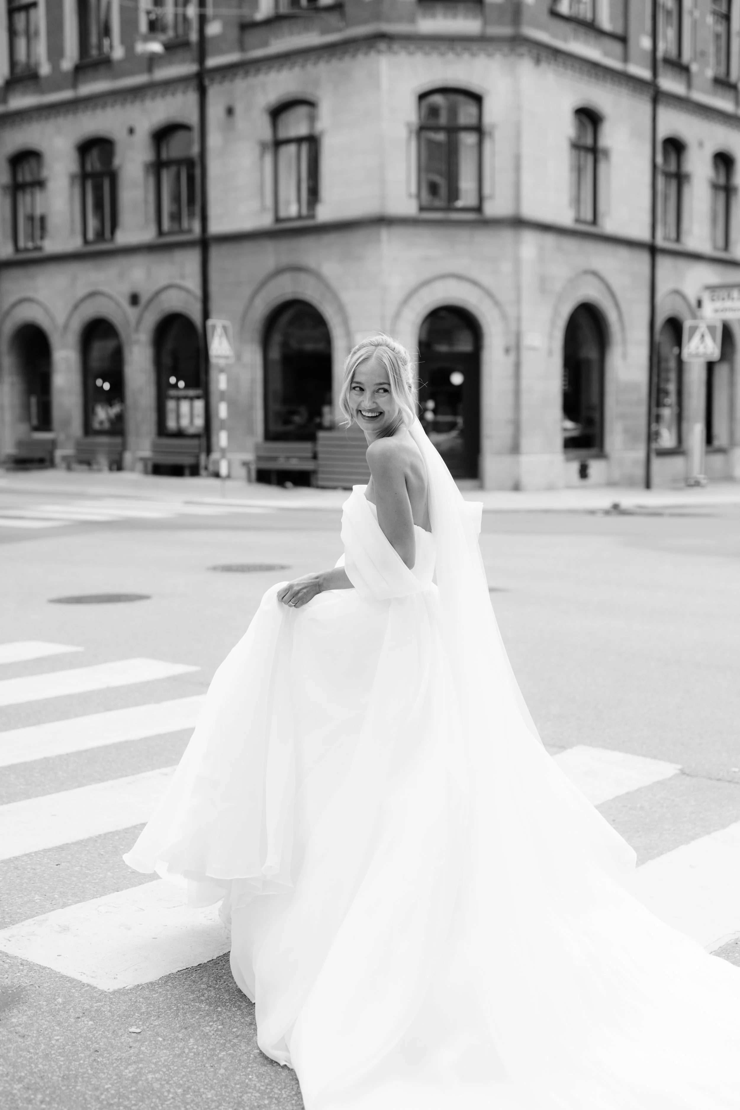 A woman in a wedding dress crossing a city street with a historical building in the background.