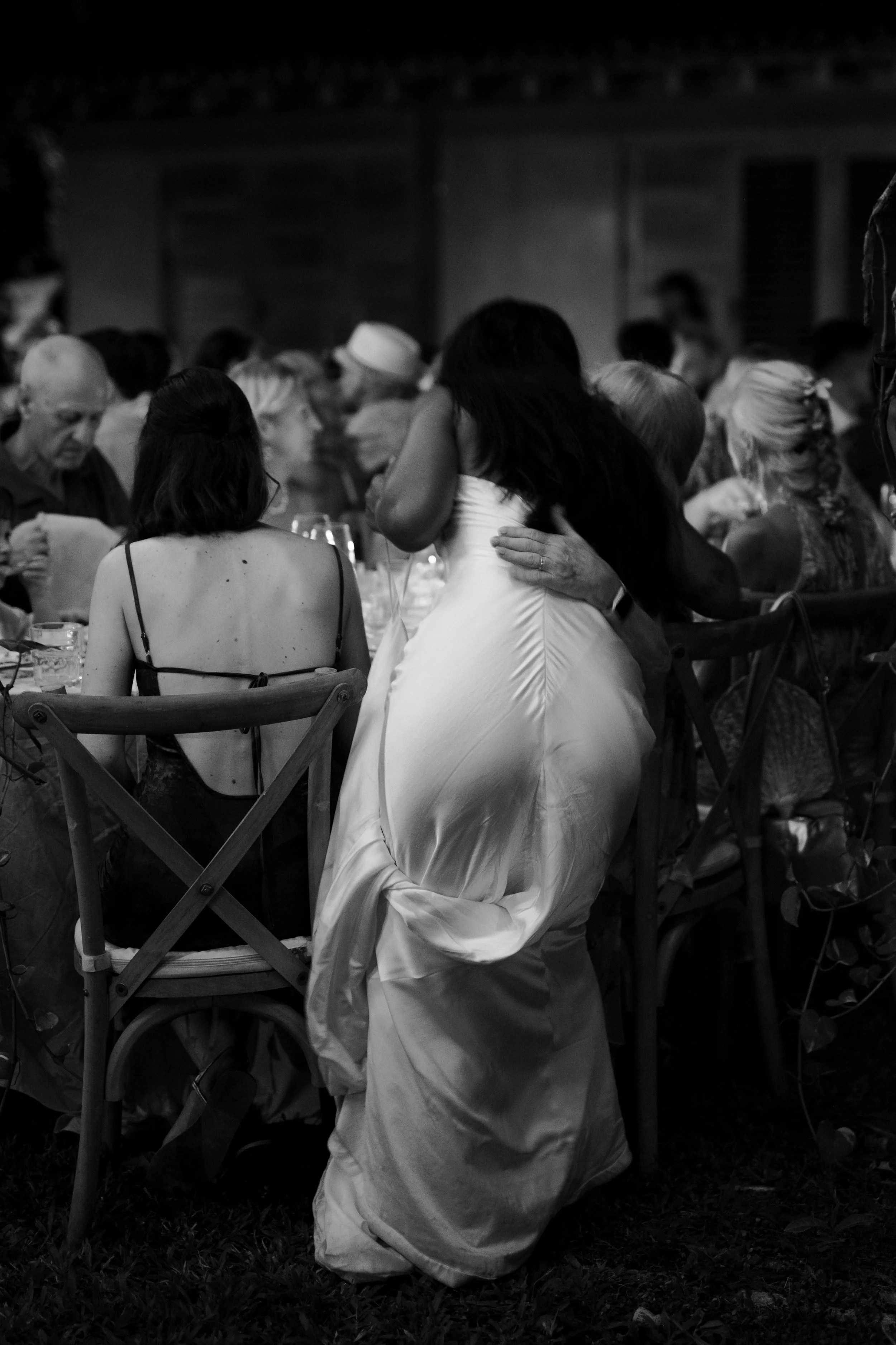 Black and white photo of women at a social gathering, one woman in a shiny dress, embracing and sitting on a man’s lap, with other people seated at a table in the background.