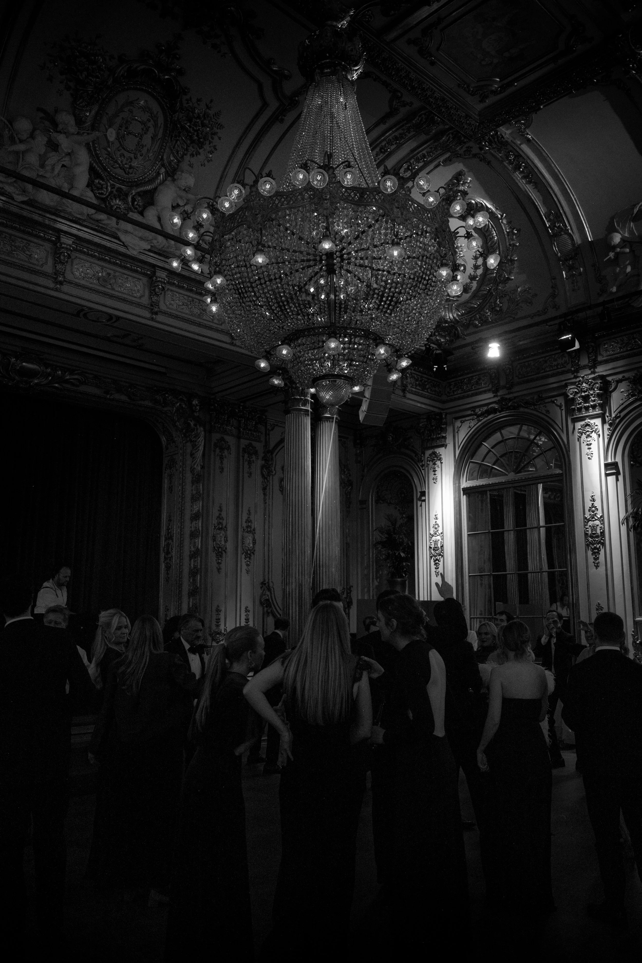 Guests dancing in a grand, ornately decorated room with a large chandelier hanging from the ceiling.