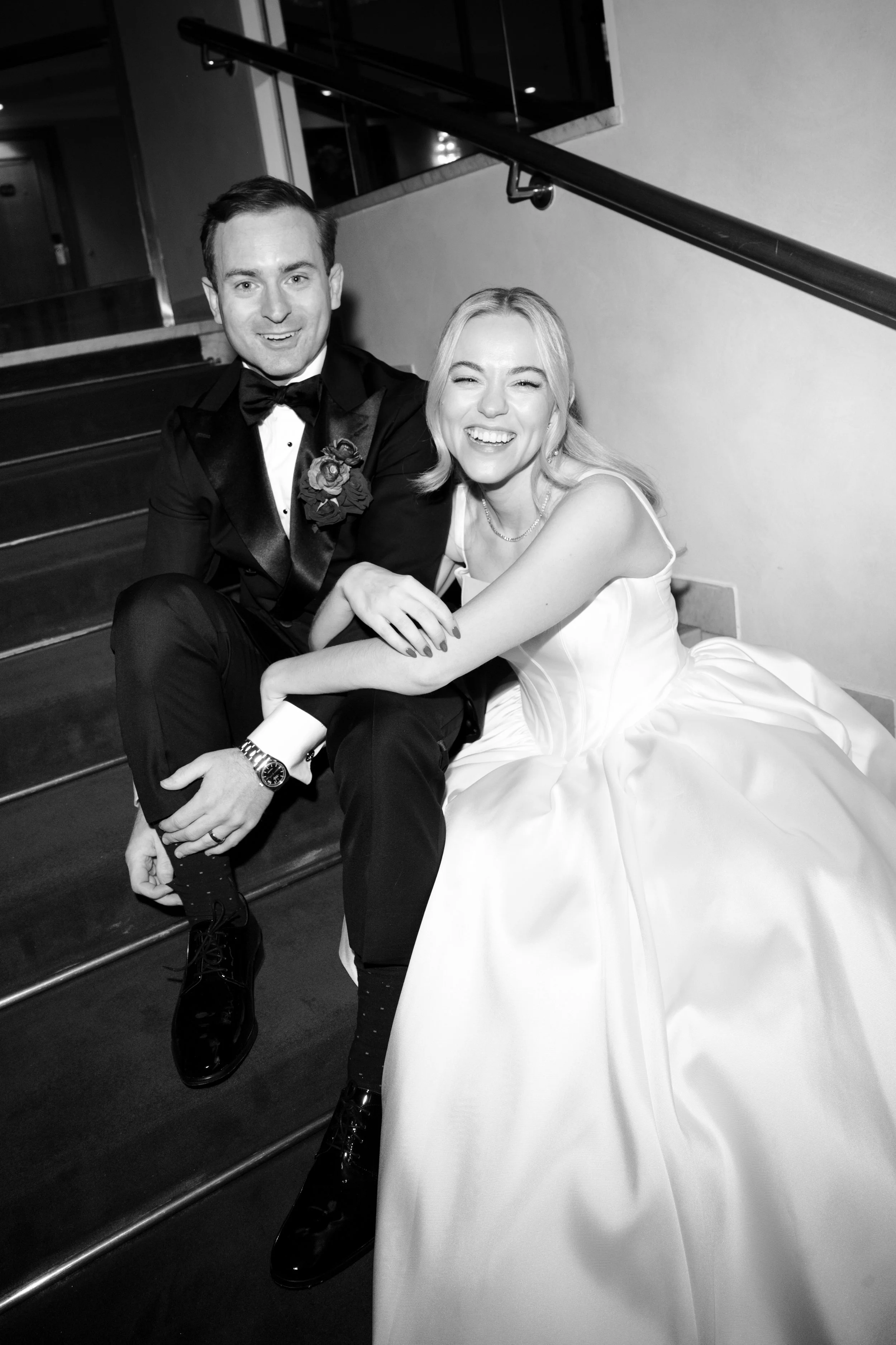 A black and white photo of a smiling bride and groom sitting on stairs at their wedding reception.