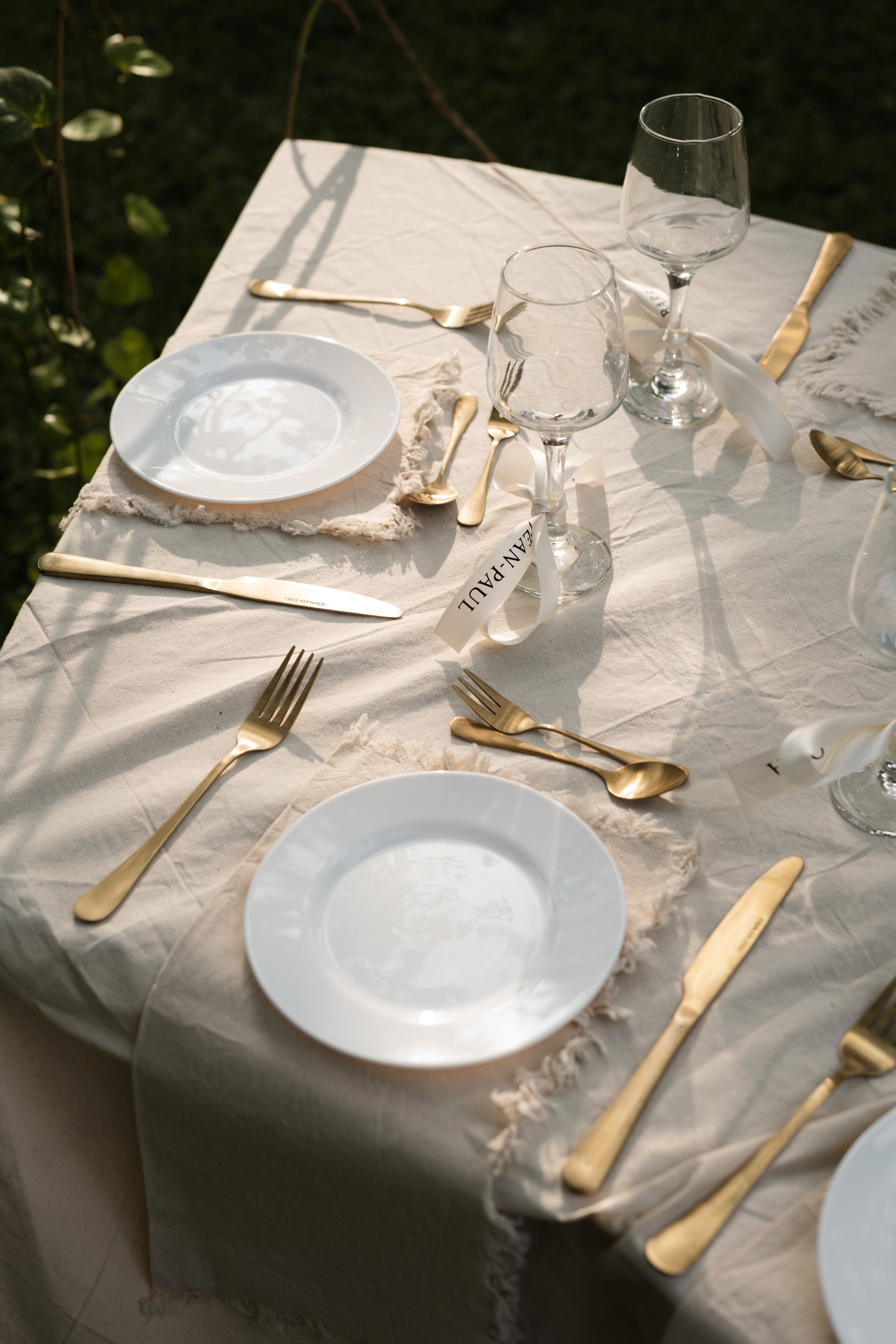 Dining table set for a meal with white plates, gold cutlery, and wine glasses, outdoors in natural lighting.