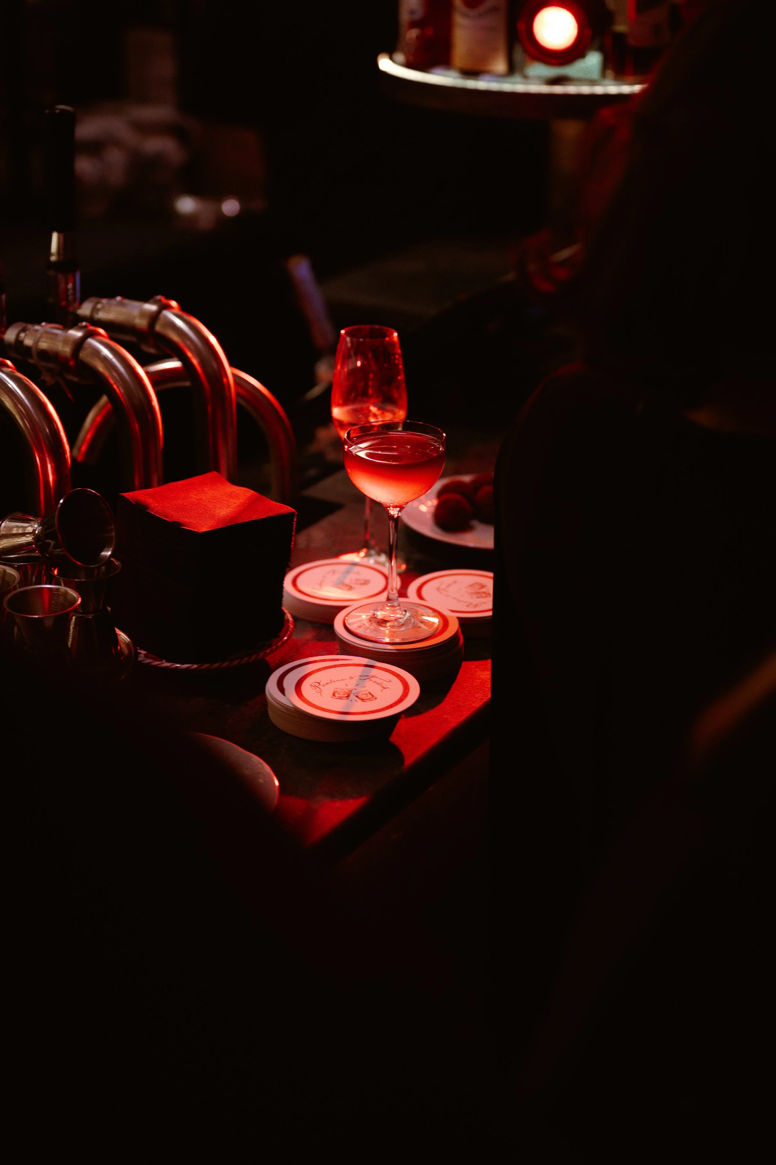 A dimly lit bar table with stacked coasters, two stemmed glasses filled with red drinks, along with napkins and bar accessories, under warm, reddish lighting.